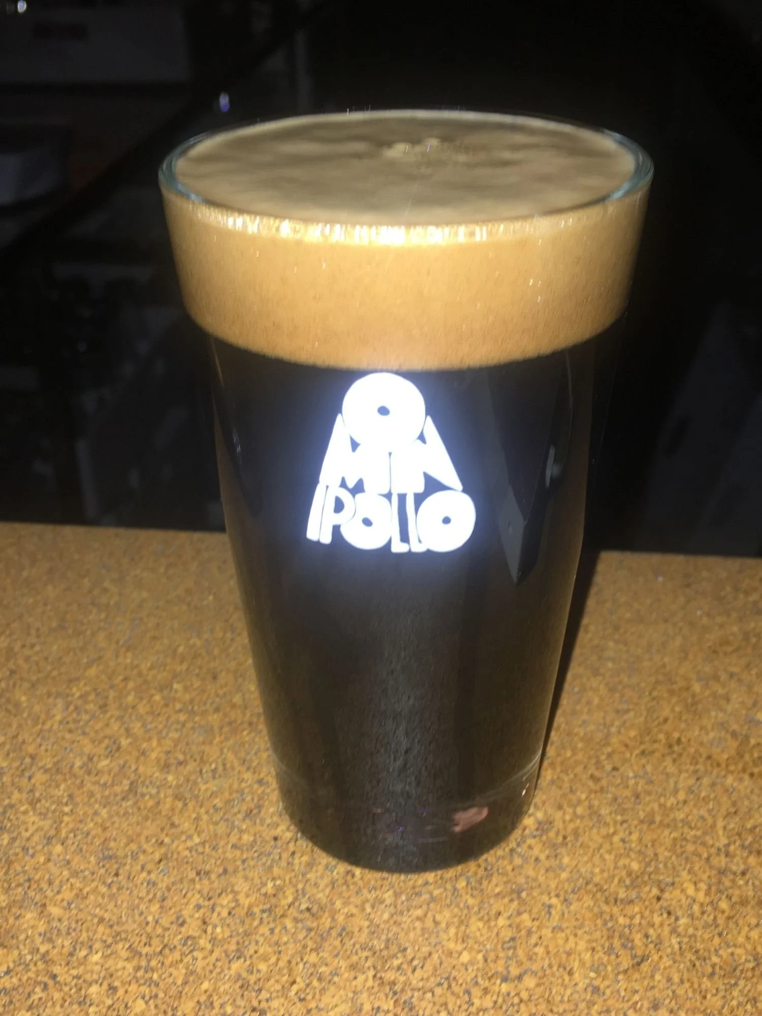 A full pint glass of dark, black imperial stout with a thick, tan head sitting on a textured brown bar surface. The glass features a prominent white logo that reads "OMNIPOLLO" across the center, set against a dark background.