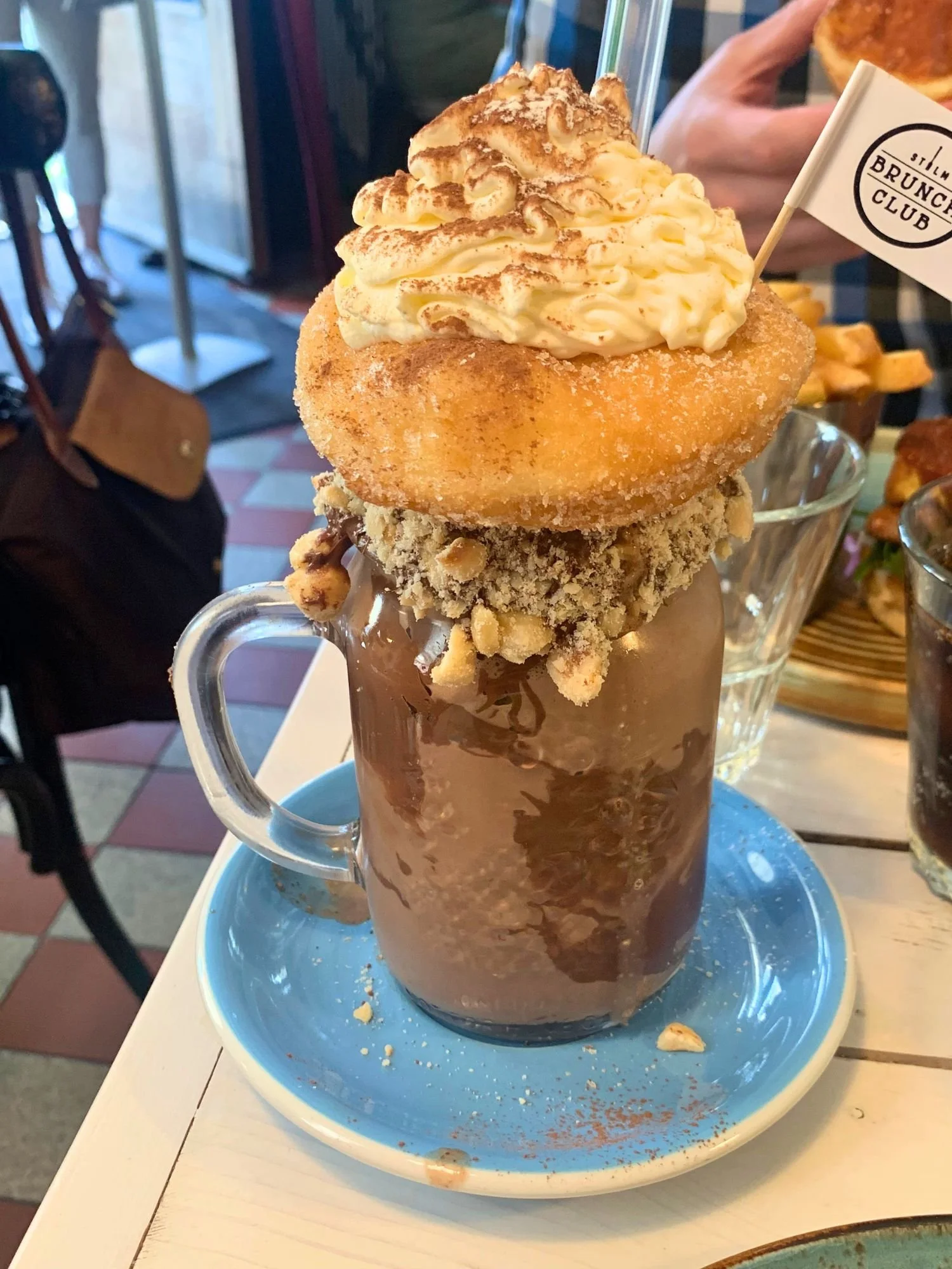 An over-the-top chocolate milkshake served in a glass mug on a light-blue saucer. The rim of the glass is coated in chocolate and crushed nuts, supporting a large sugar-dusted donut balanced on top.