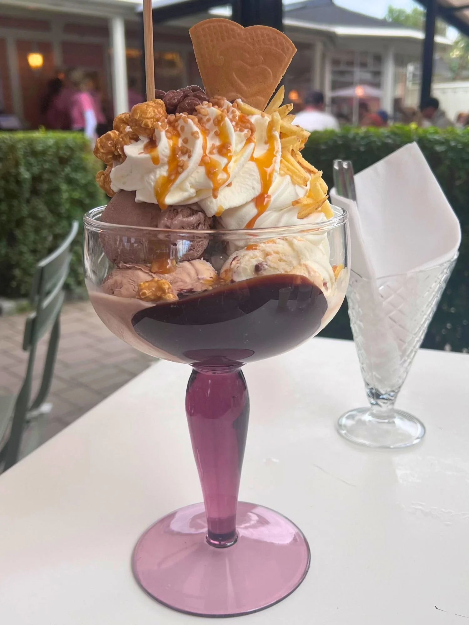An elaborate chocolate and caramel ice cream sundae served in a purple-stemmed glass goblet on an outdoor cafe table.