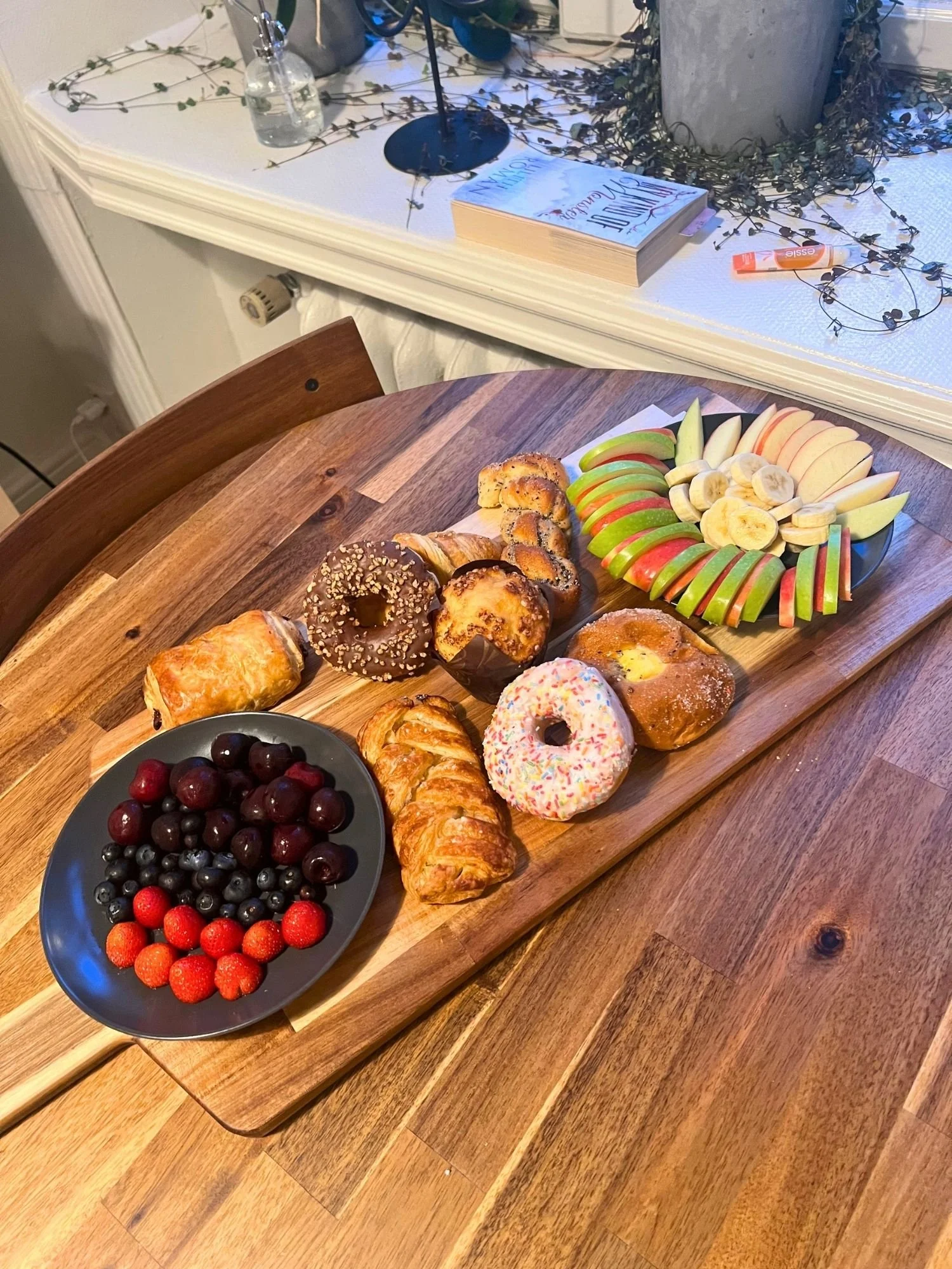 A long wooden serving board rests diagonally across a round wooden table, featuring an assortment of pastries, donuts, and fruit.