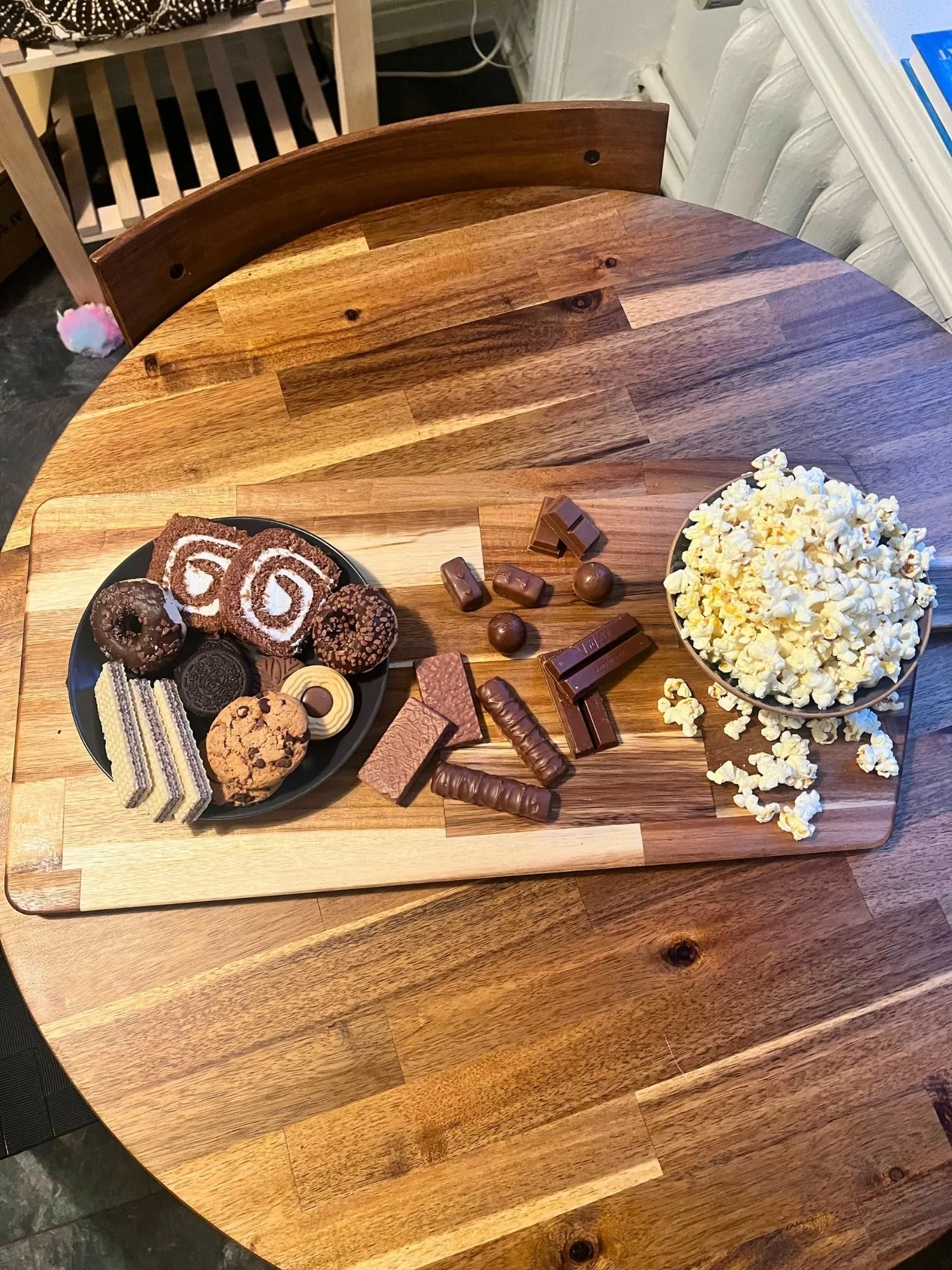 High-angle, top-down shot of a wooden serving board set on a round wooden table, holding a plate of cookies and mini donuts, loose chocolate candy bars, and a bowl of popcorn.