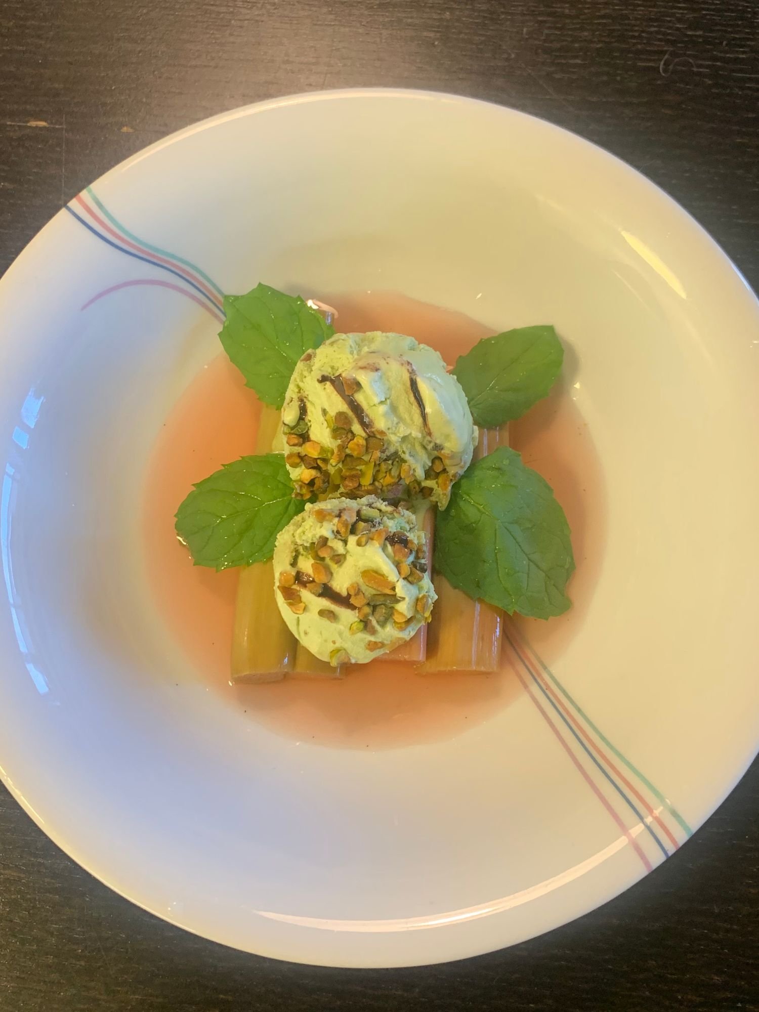 Pistachio ice cream scoops over cooked rhubarb stalks in a pink syrup, garnished with mint leaves in a white bowl.