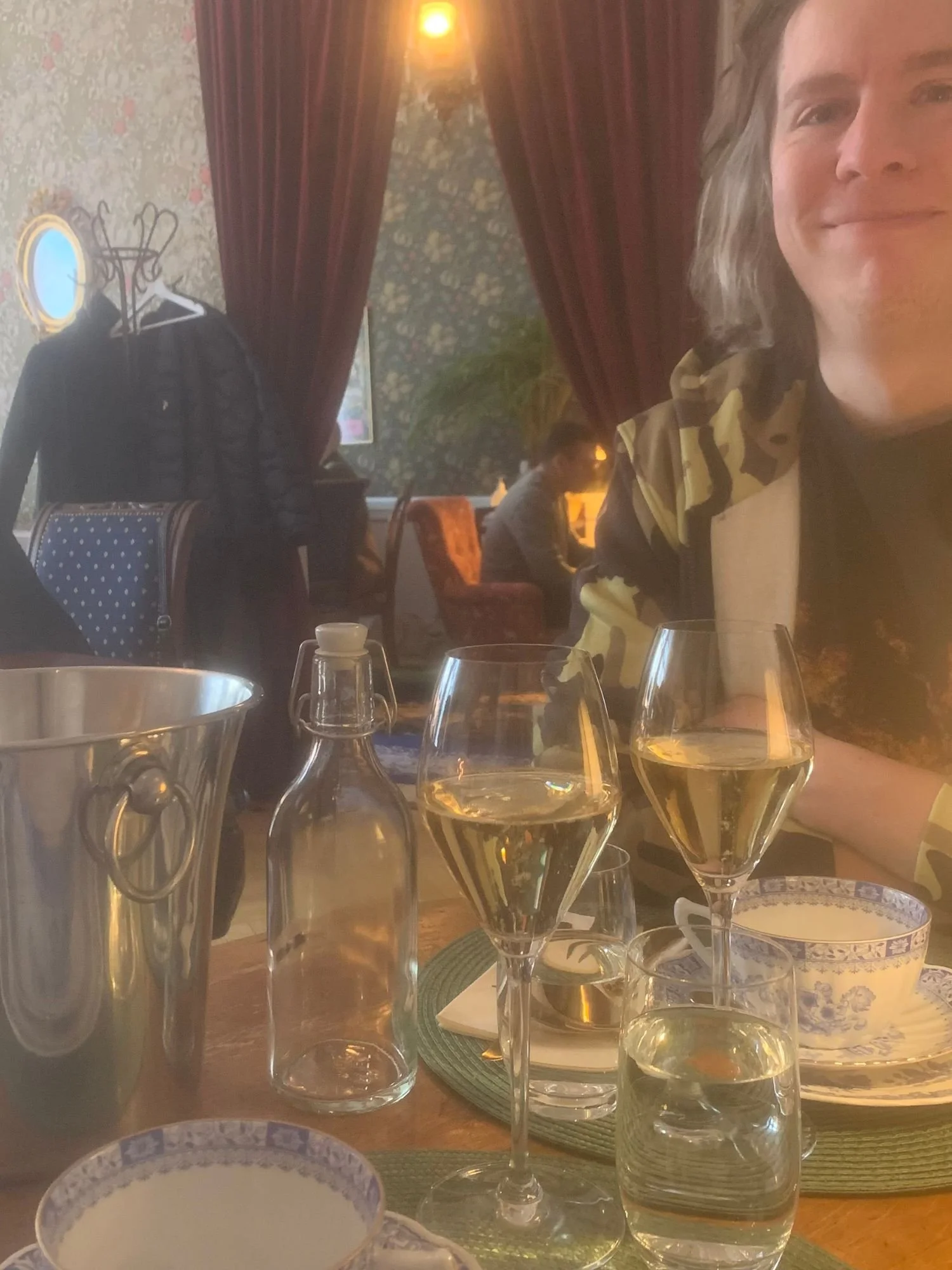 A man sitting at a table at Alice och Kattens Tesalong in Lund. Two glasses filled with champagne, a clear swing-top glass bottle, and a glass of water.
