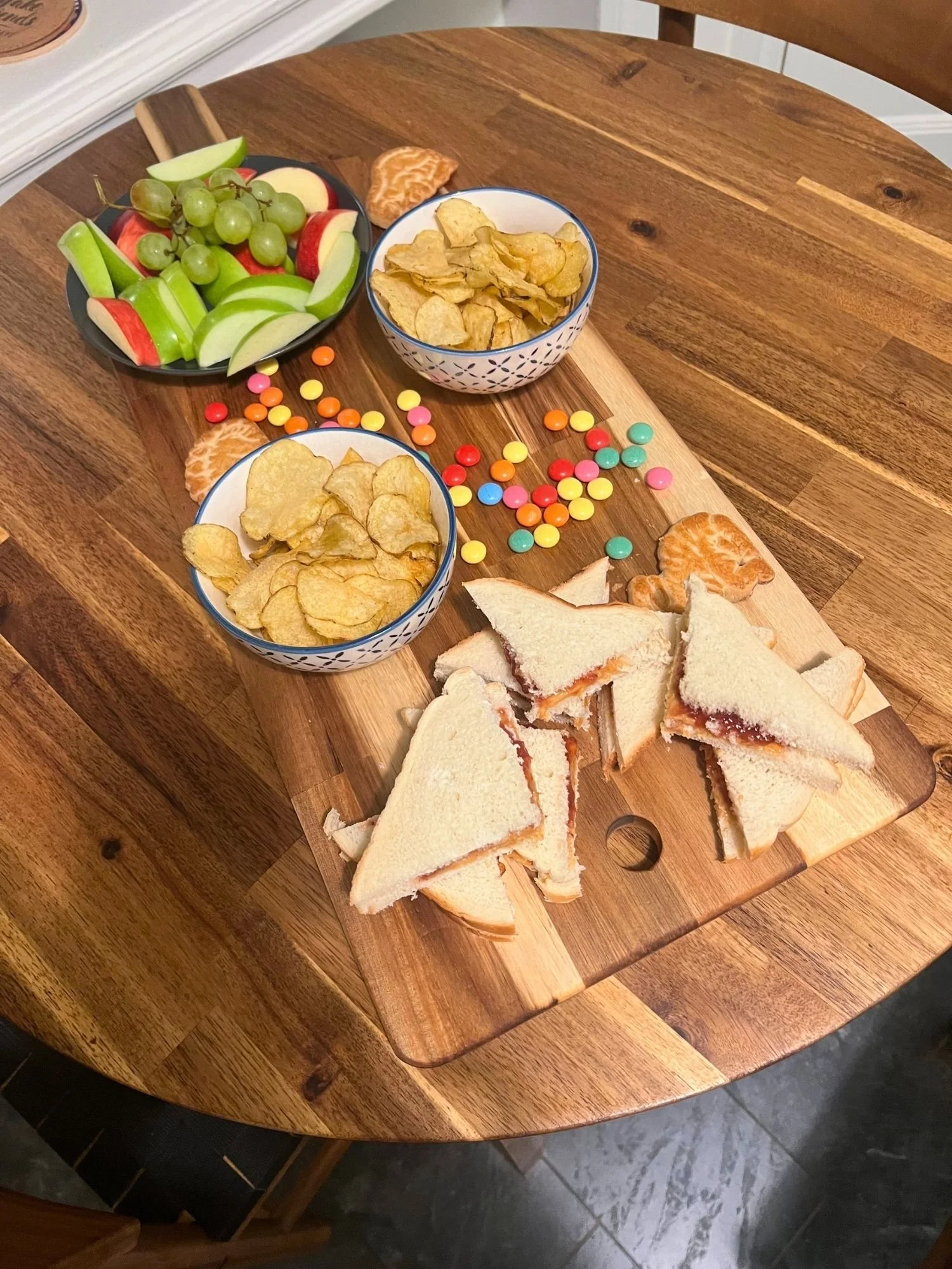 High-angle shot of a rectangular wooden serving board on a round wooden table, featuring a kid-friendly spread of cut sandwich triangles.