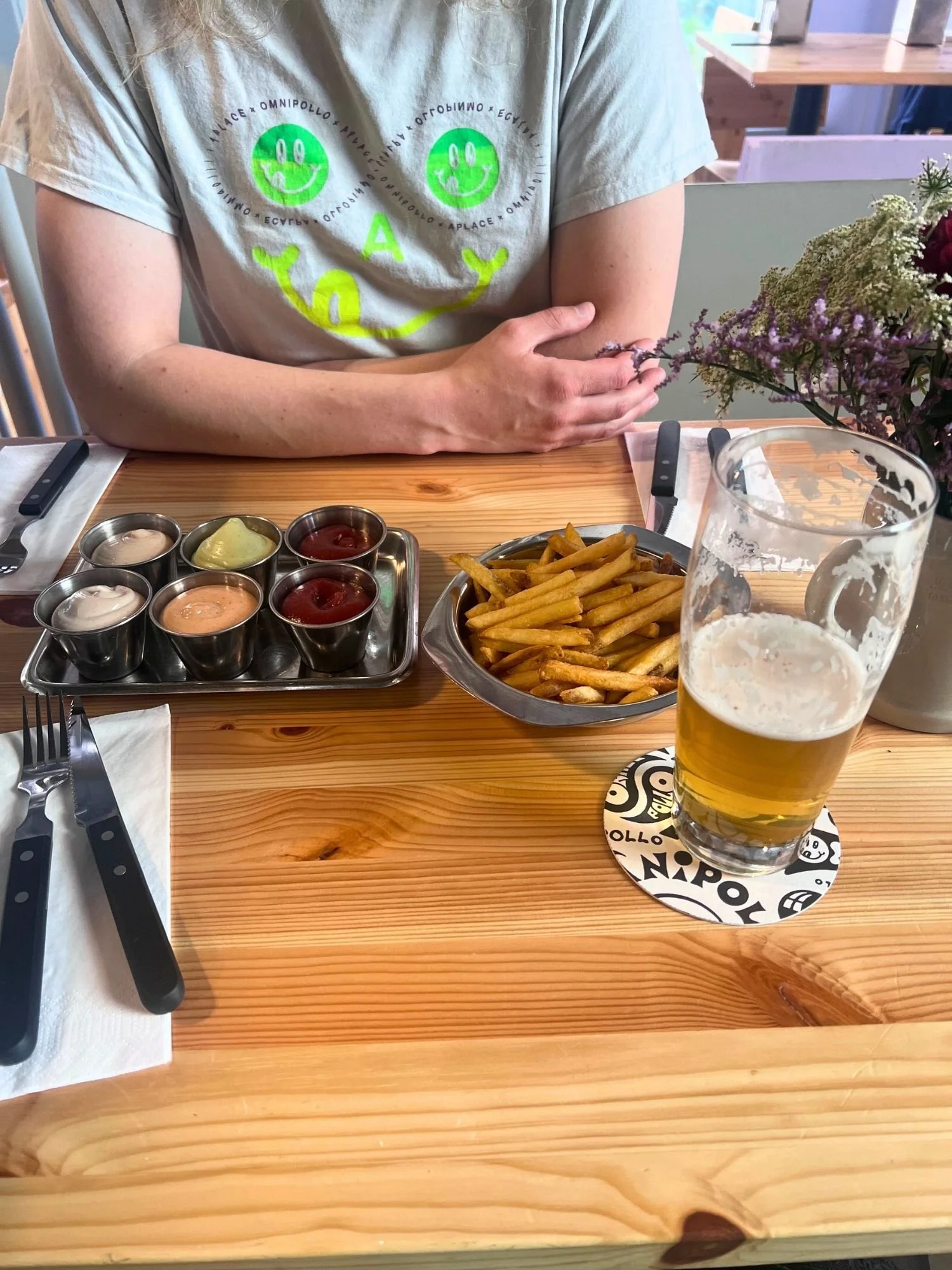 A table spread featuring a metal bowl of french fries, a tray holding six small metal cups of various dipping sauces, and a glass of golden beer resting on an Omnipollo coaster.