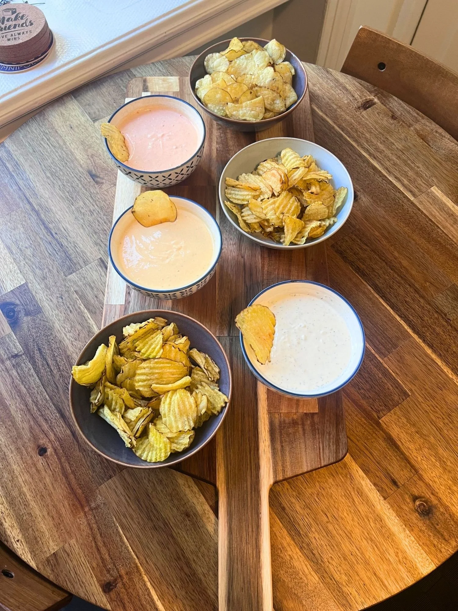High-angle, length-wise shot of six ceramic bowls filled with potato chips and savory dips arranged on a wooden serving board on a round wooden table.