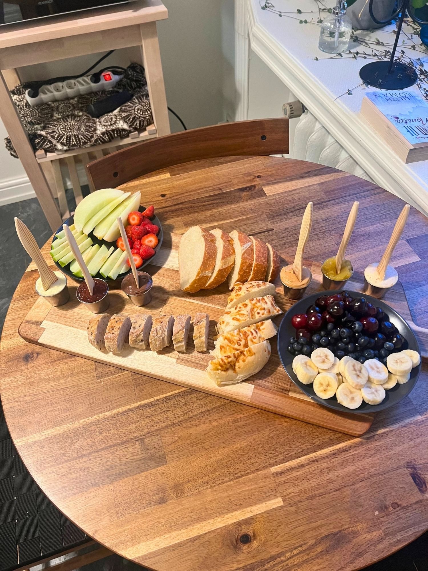 A wooden serving board on a round wooden table, arranged with sliced artisan breads, plates of fresh fruit, and small ramekins of spreads with wooden knives.