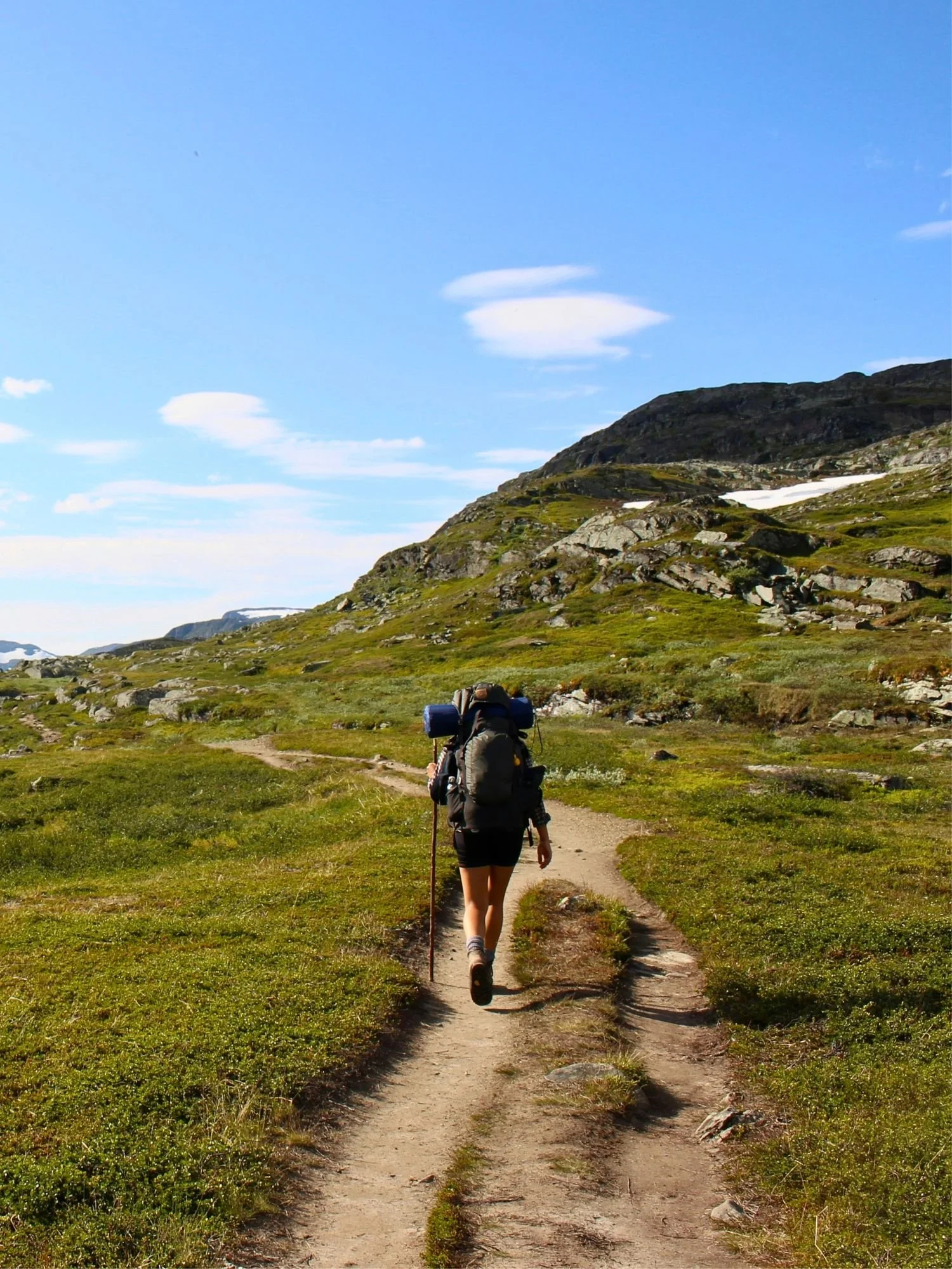 A backpacker walking along a narrow, dirt trail through a vast mountain landscape. The hiker wears a large, dark grey trekking pack with a blue sleeping mat strapped to the top and carries a wooden walking stick.