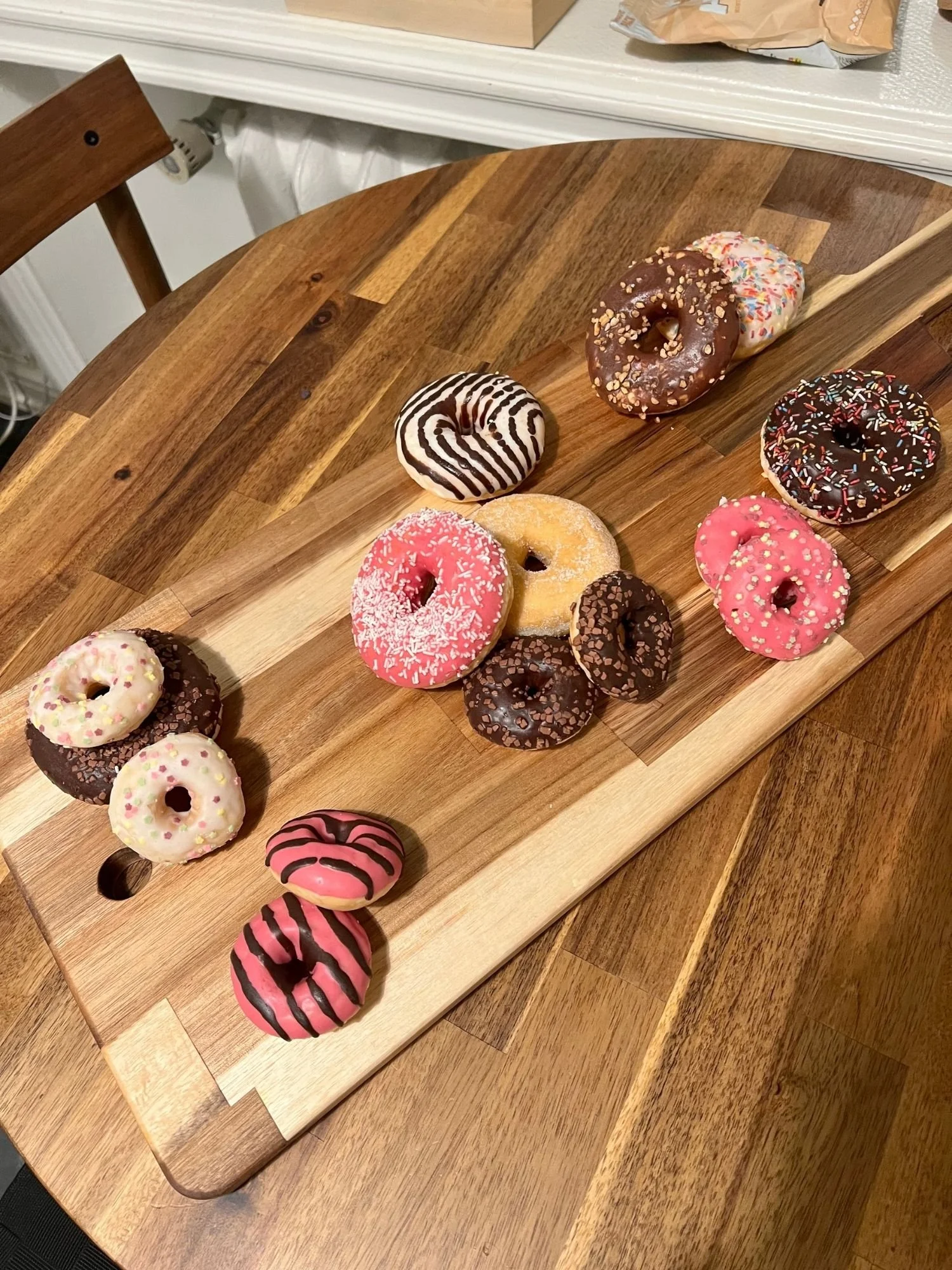 A long wooden serving board rests diagonally across a round wooden table, displayed with an assortment of colorful iced donuts.