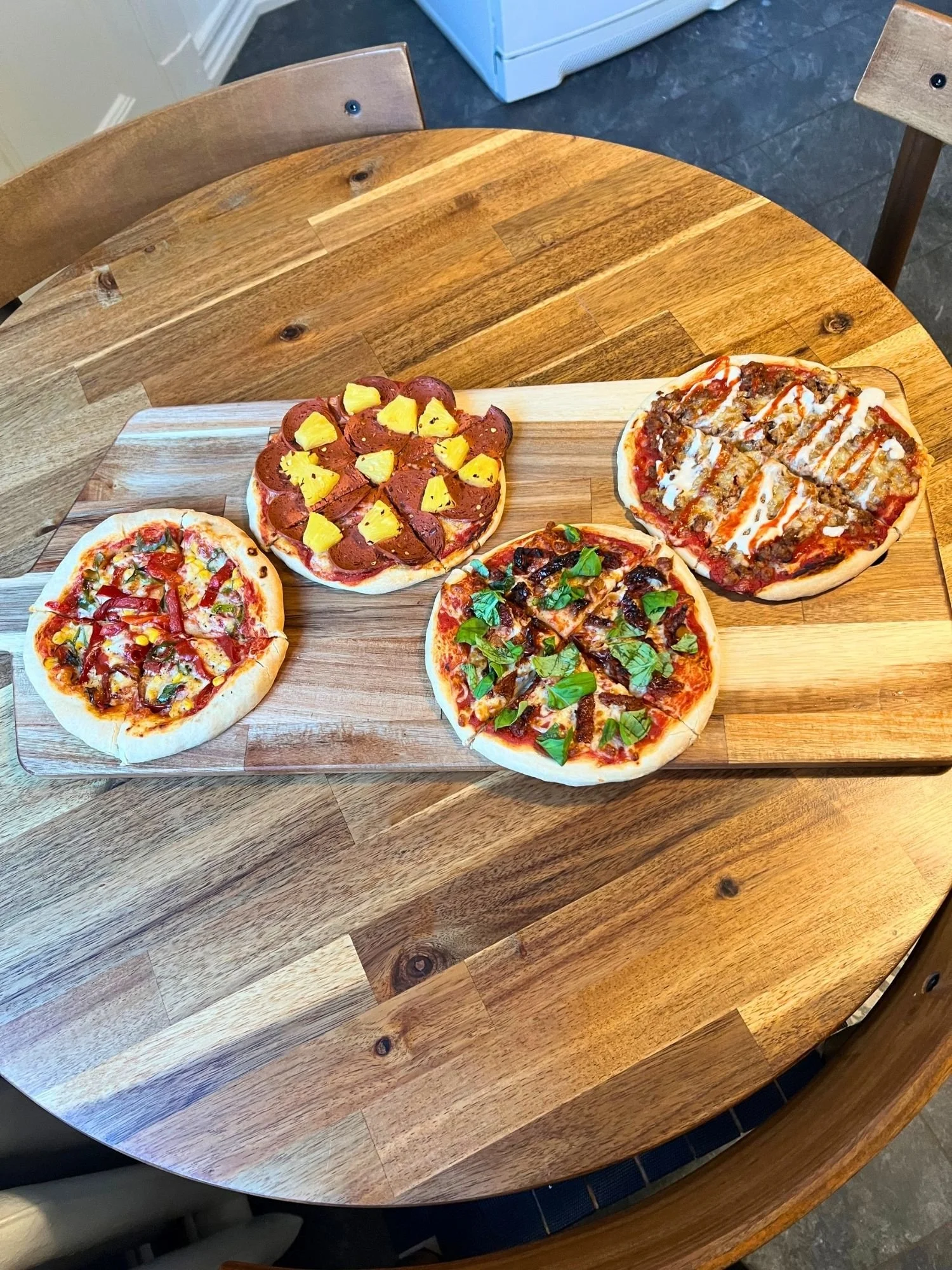 Four personal-sized mini pizzas arranged on a large rectangular wooden serving board placed on a round wooden table.