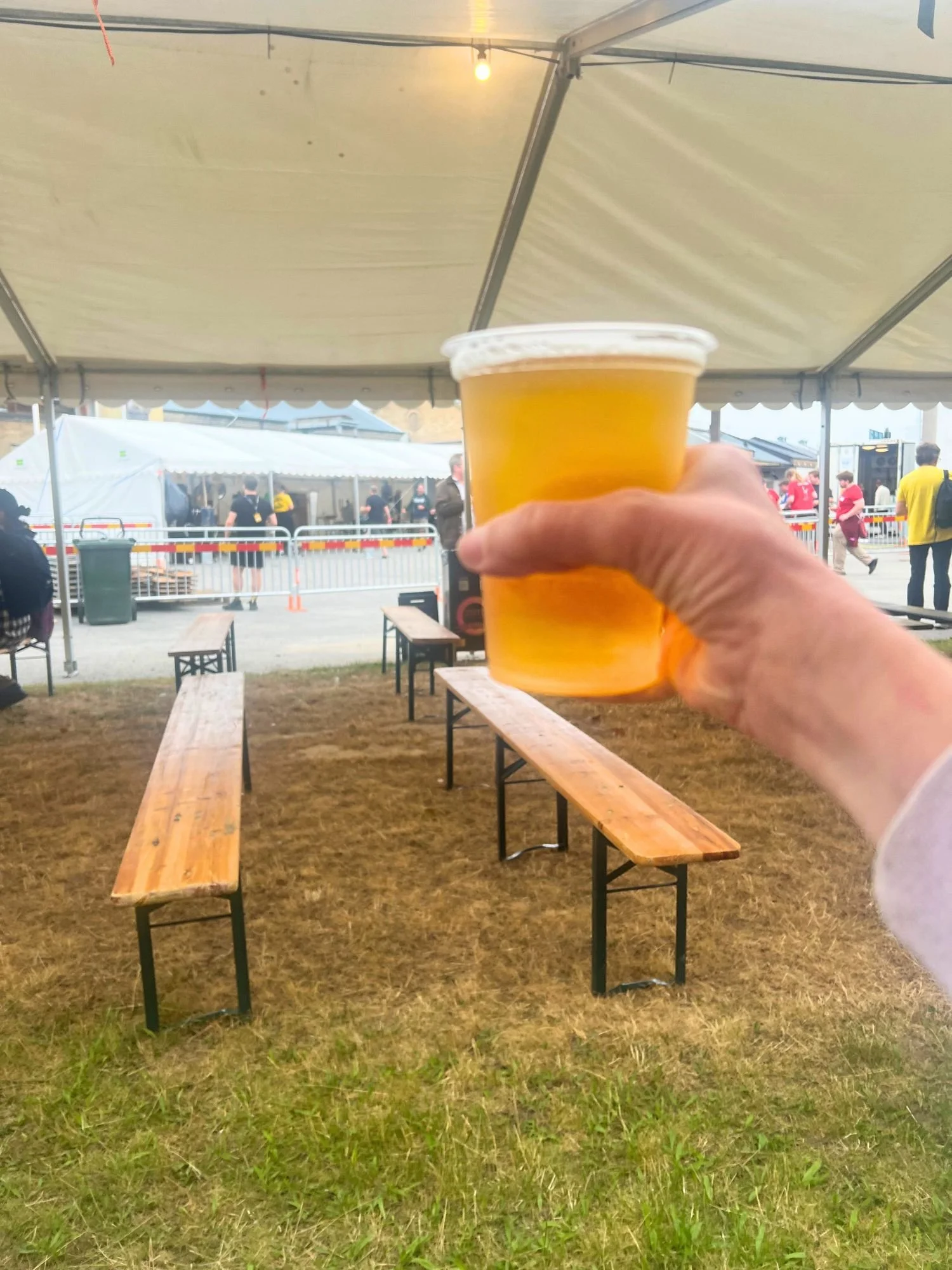 A person holds a clear plastic cup of golden beer at an outdoor beer festival, with tents, picnic benches, and festival-goers in the background.