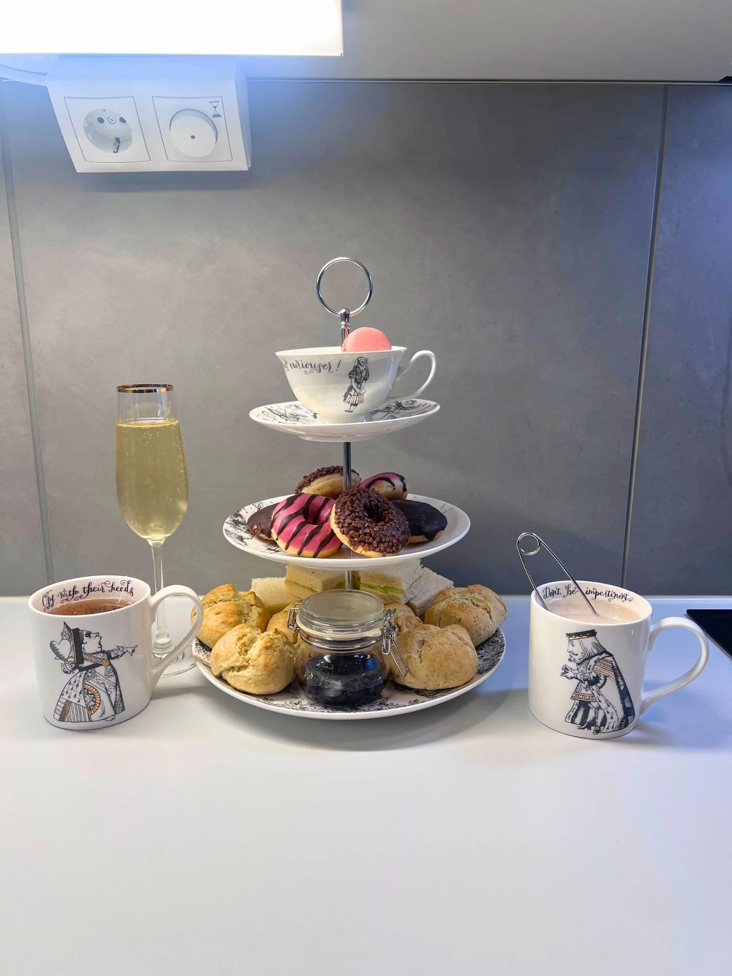 A three-tiered tea stand filled with sweets and savories on a kitchen counter. The top tier has a white teacup with an Alice in Wonderland illustration on a matching saucer, holding a single pink macaron.