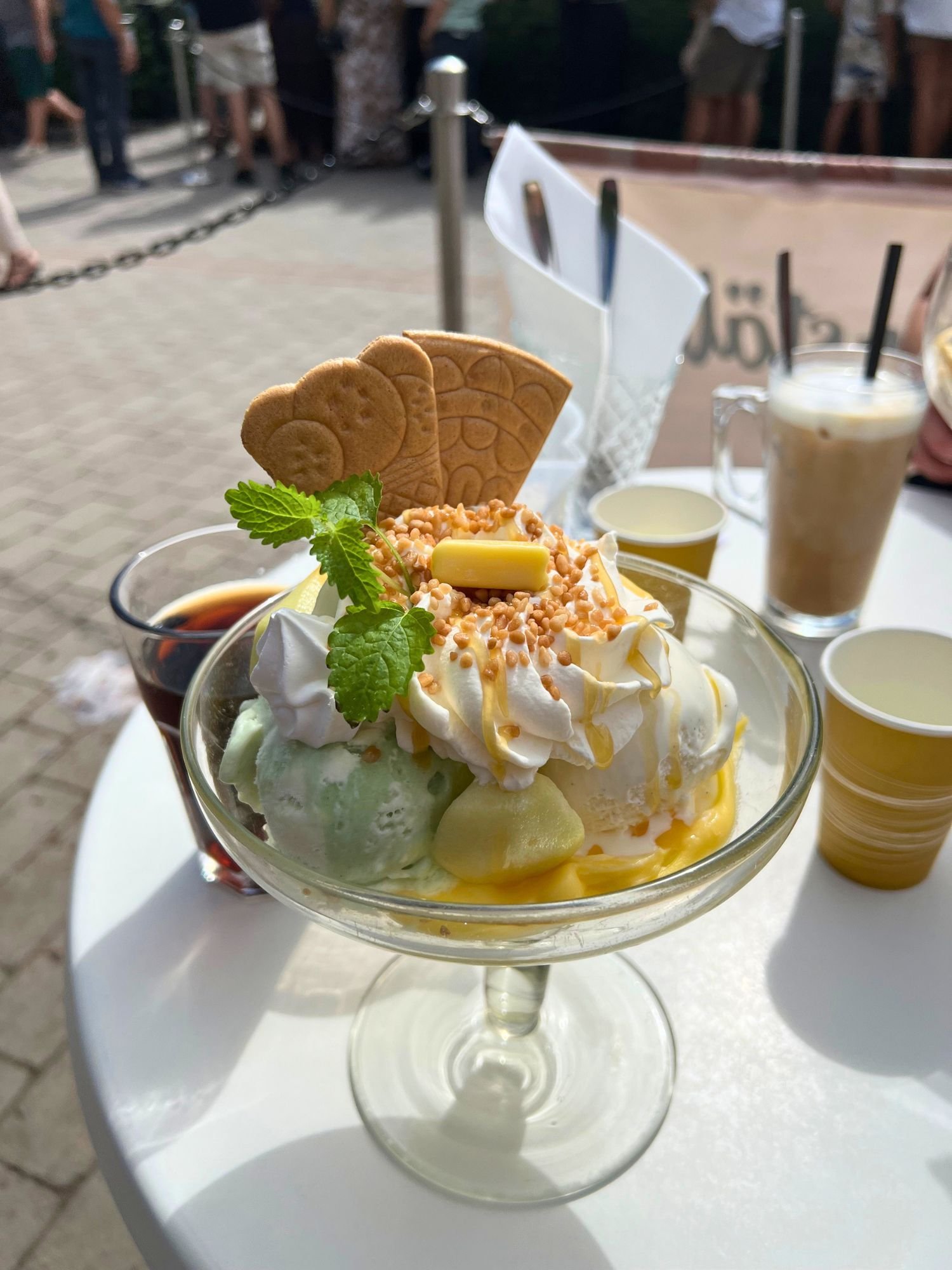 An elegant ice cream sundae served in a large, clear-stemmed glass bowl on a white outdoor table.