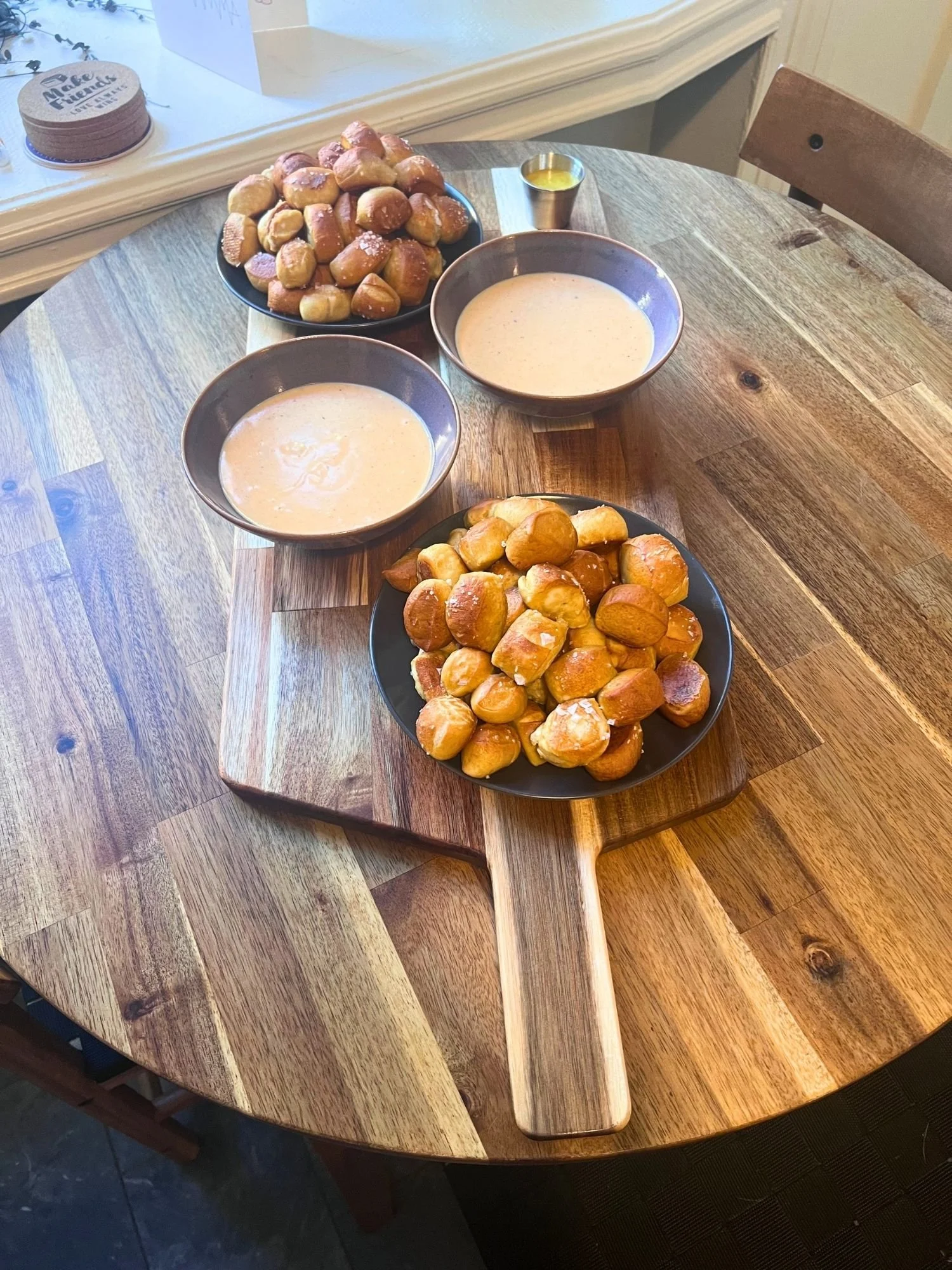 High-angle, length-wise view of a wooden serving paddle on a round wooden table, arranged with two plates of pretzel bites, two bowls of cheese dip, and a small metal cup of mustard sauce.