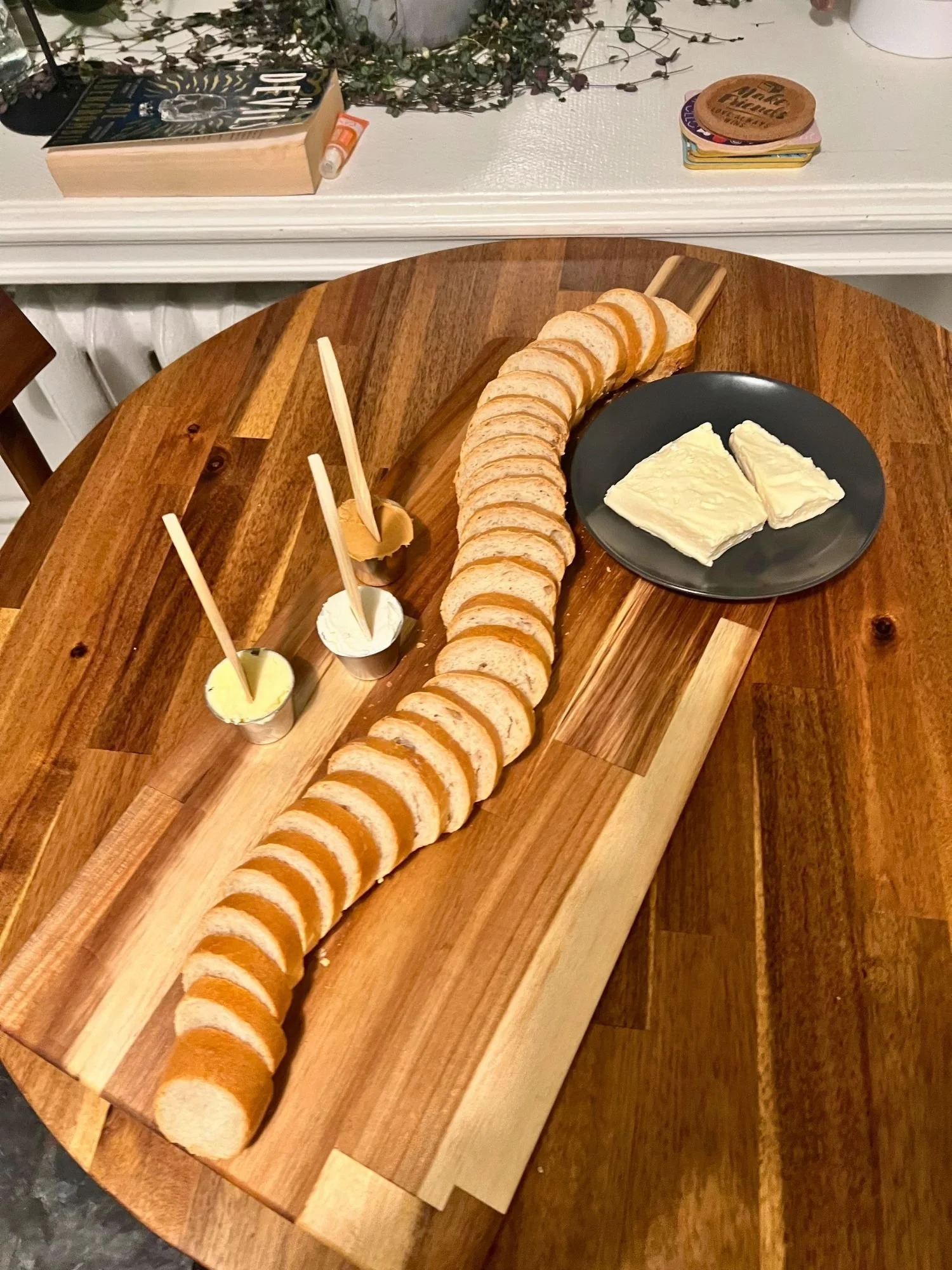 A long wooden serving board rests diagonally across a round wooden table, presenting a sliced baguette arranged in a long S-curve down its length.