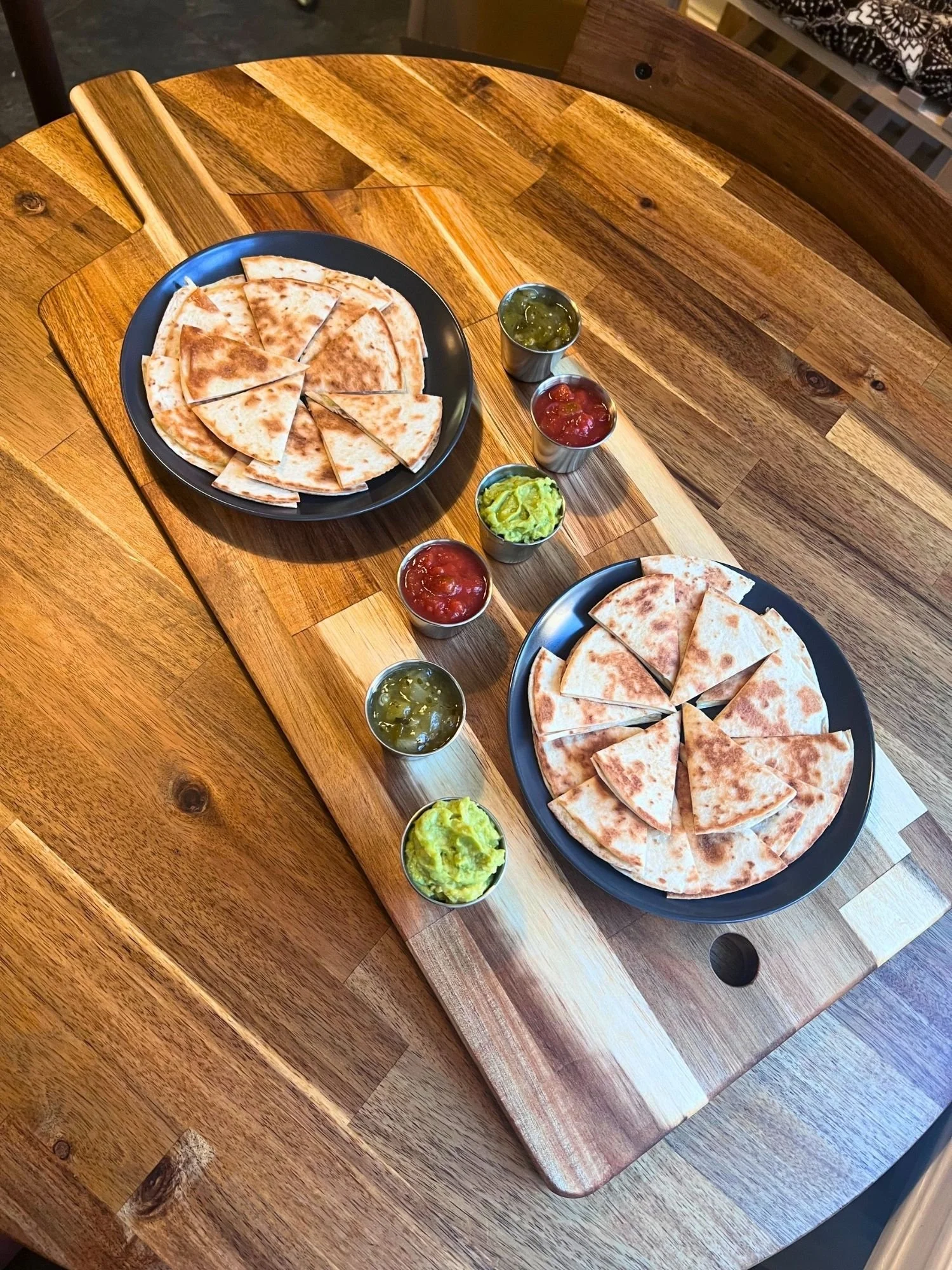 A large wooden paddle board on a round wooden table set with a spread of quesadillas and dips.