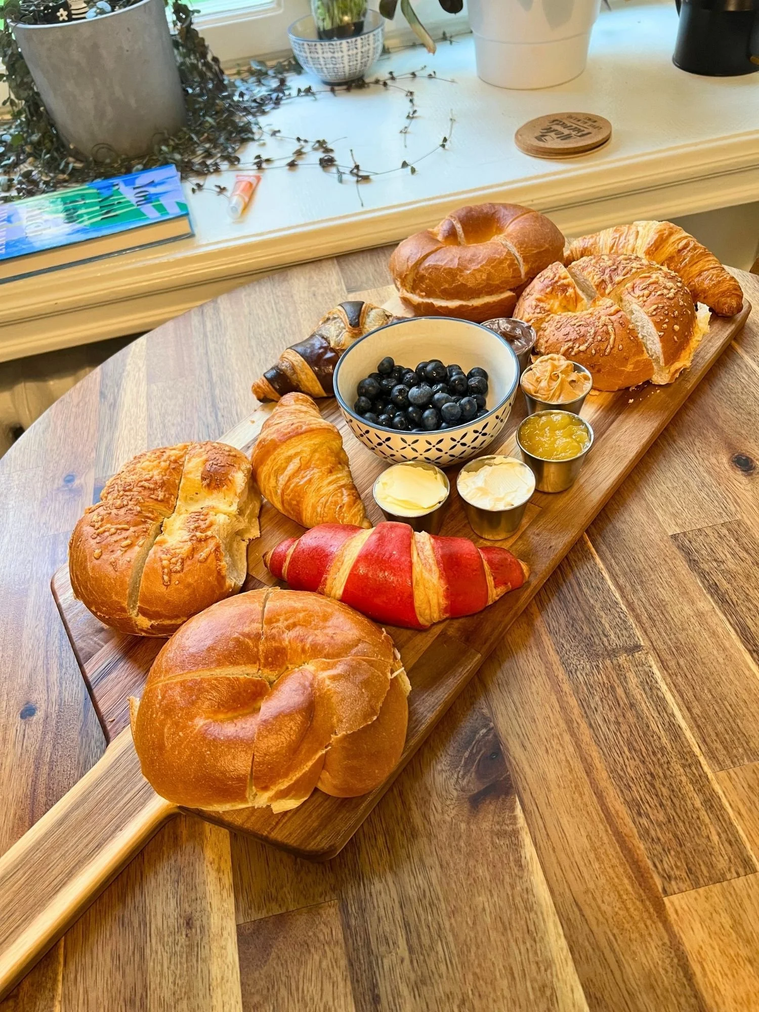 A long wooden serving board with a handle stretches diagonally across a wooden table, presenting a breakfast board of bagels, croissants, fresh blueberries, and spreads.