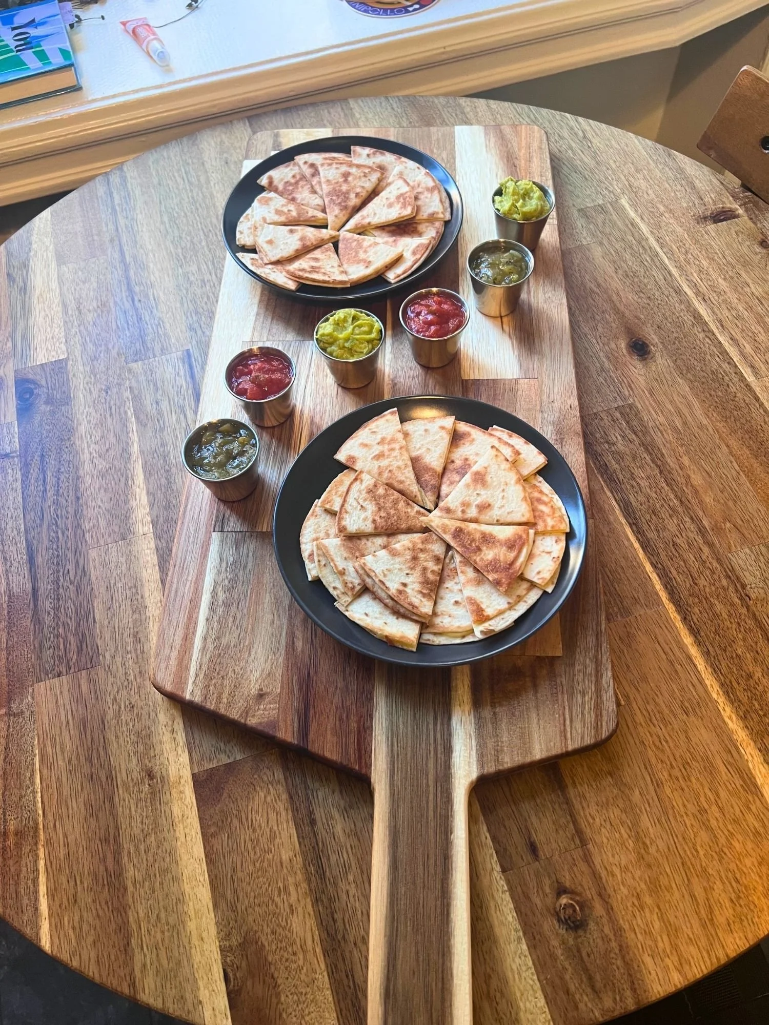A long wooden serving paddle rests vertically on a round wooden table, carrying a quesadilla spread.