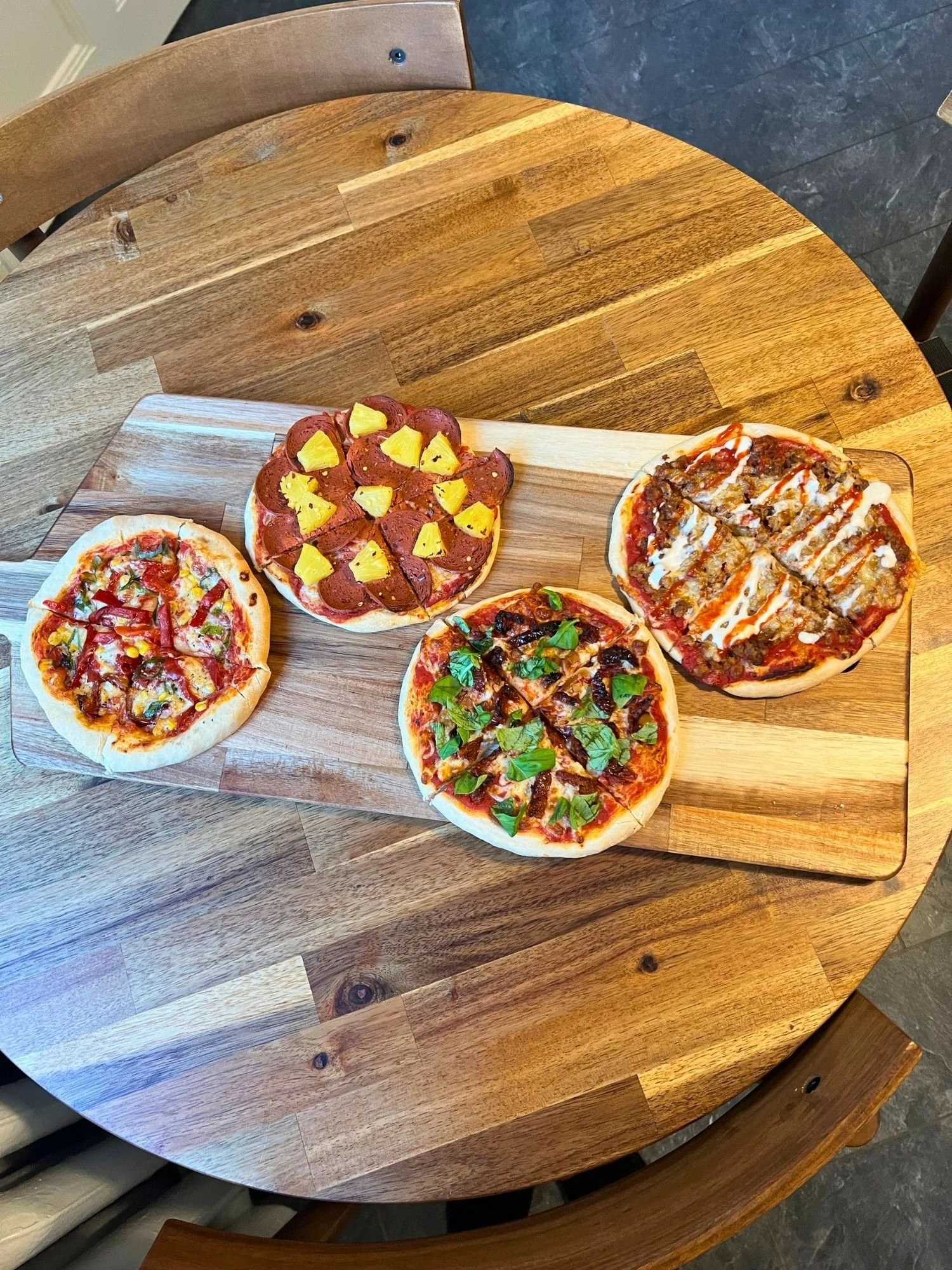High-angle shot of a rectangular wooden serving board on a round wooden table, set with four small personal-sized pizzas featuring varied toppings.