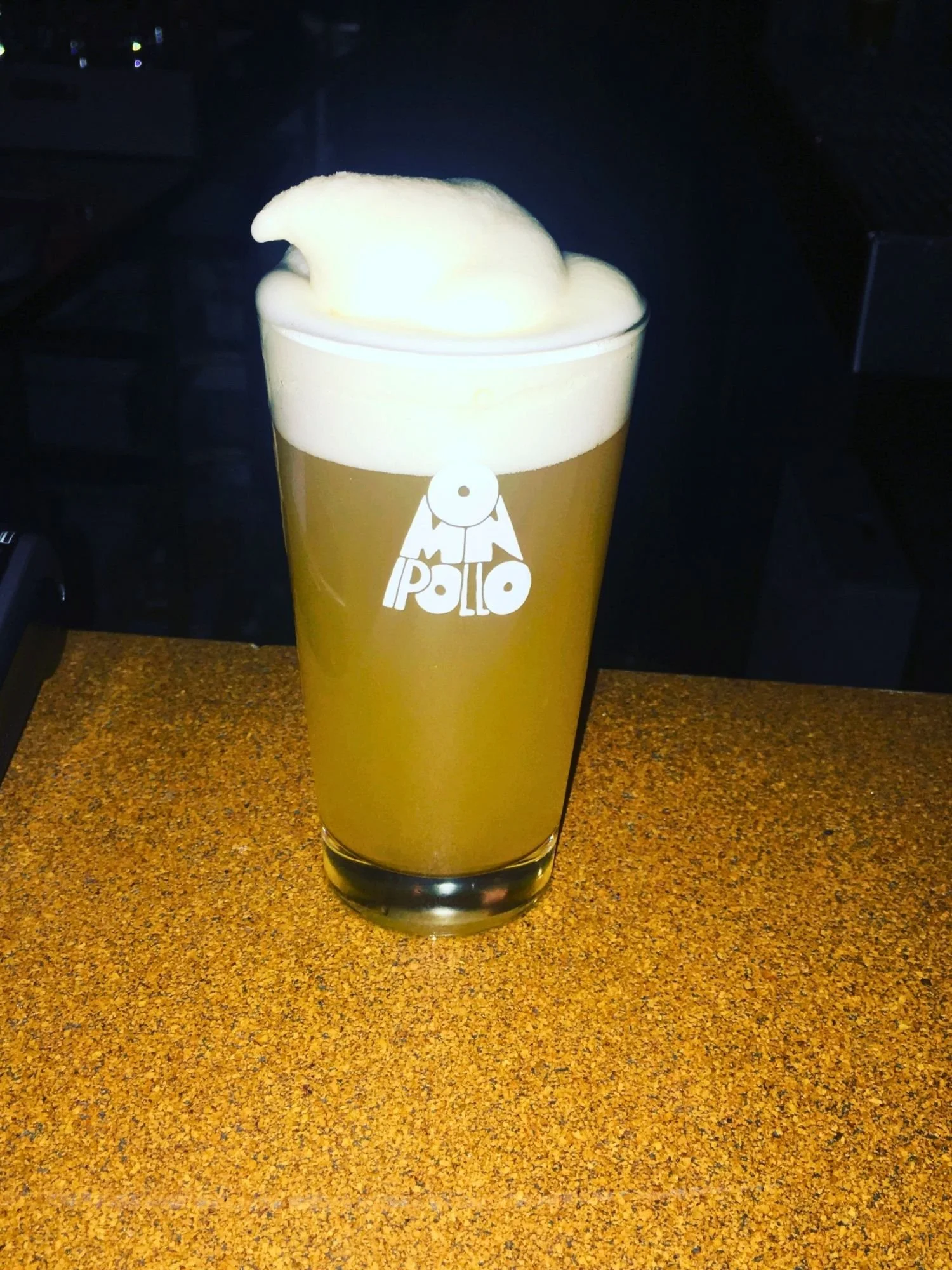 A glass of hazy, golden-yellow beer on a cork countertop. The pint glass features a white logo that says "OMNIPOLLO". It's topped with a thick foam crown that swirls up into a point at the top.
