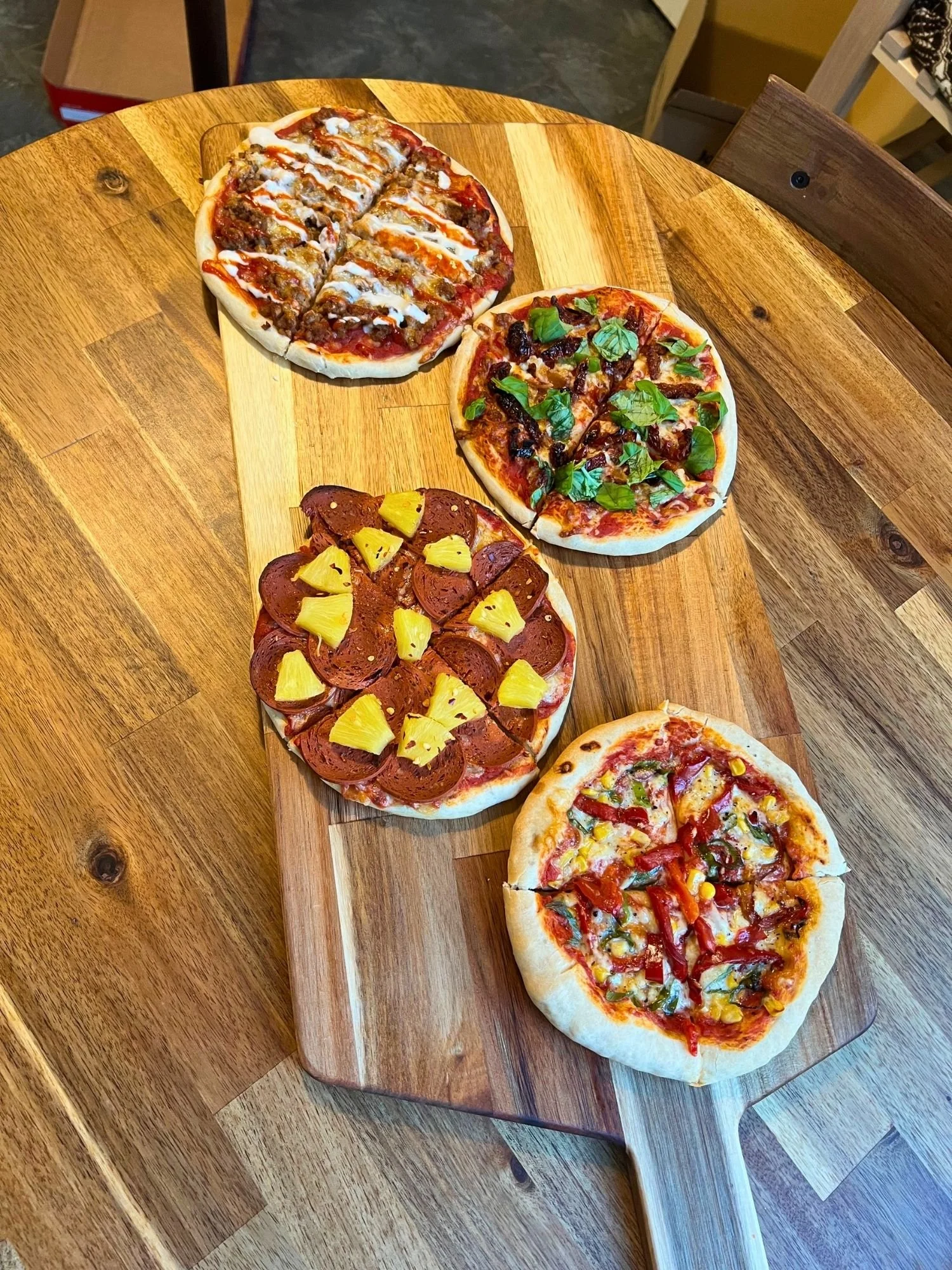 A long wooden paddle-style board lies vertically on a round wooden table, displaying four small personal-sized pizzas.