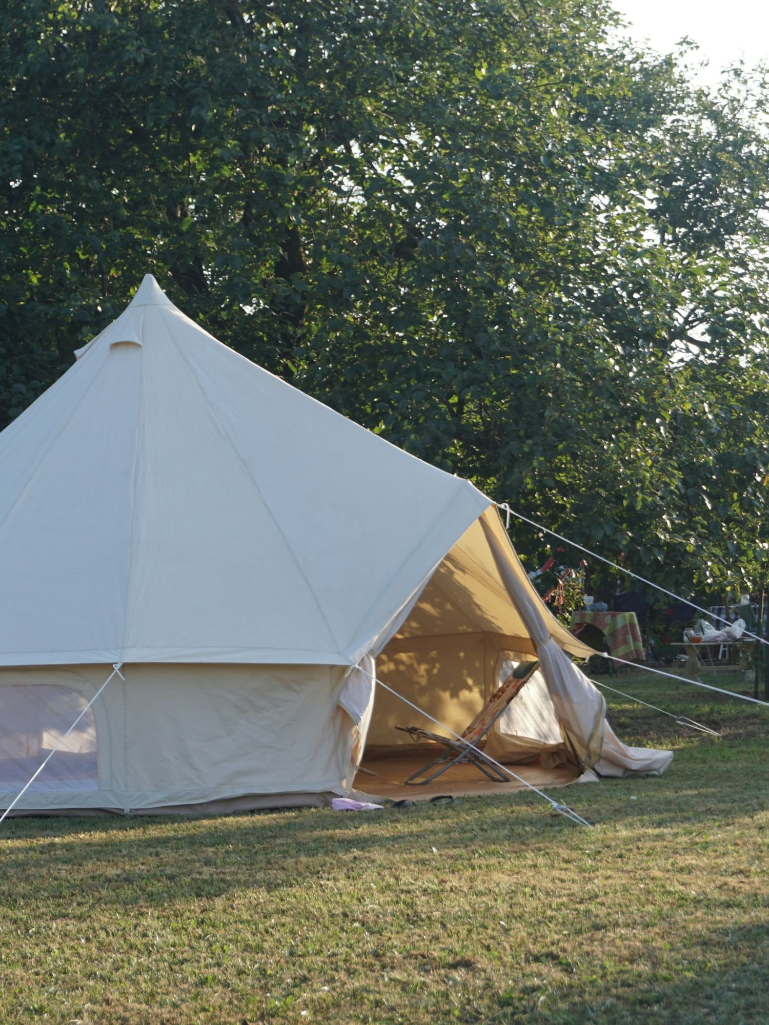 A large, cream-colored canvas bell tent is pitched on a grassy lawn with a thick line of green trees in the background. The tent's large triangular entrance is wide open, revealing a tan interior where a folded patterned lounge chair rests.