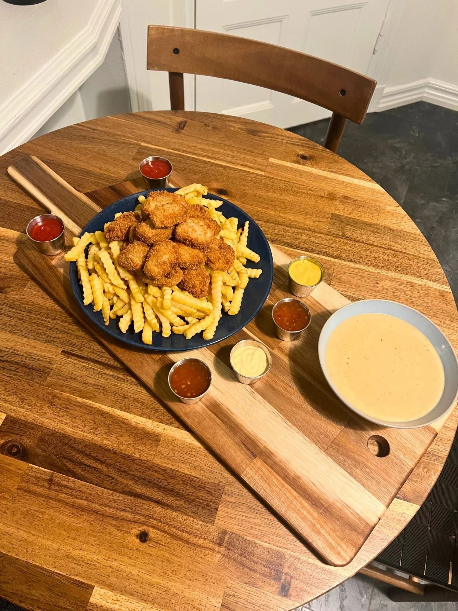 High-angle shot of a rectangular wooden serving board on a round wooden table, featuring a large blue plate piled high with crinkle-cut fries and breaded nuggets, surrounded by multiple small dipping sauces and a large bowl of cheese dip.