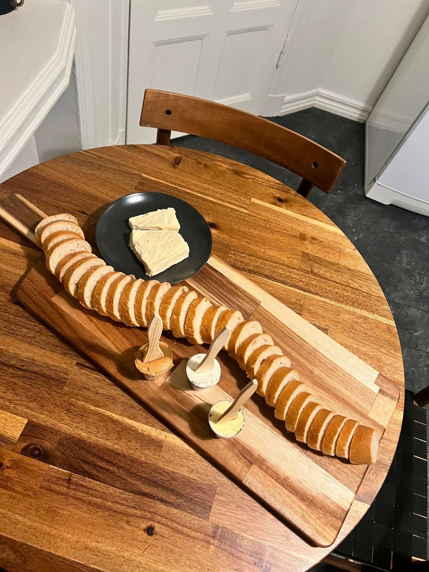 High-angle shot of a long wooden serving board on a round wooden table, featuring a sliced baguette, a small black plate with cheese slices, and three small ramekins of spreads with wooden wooden knives.