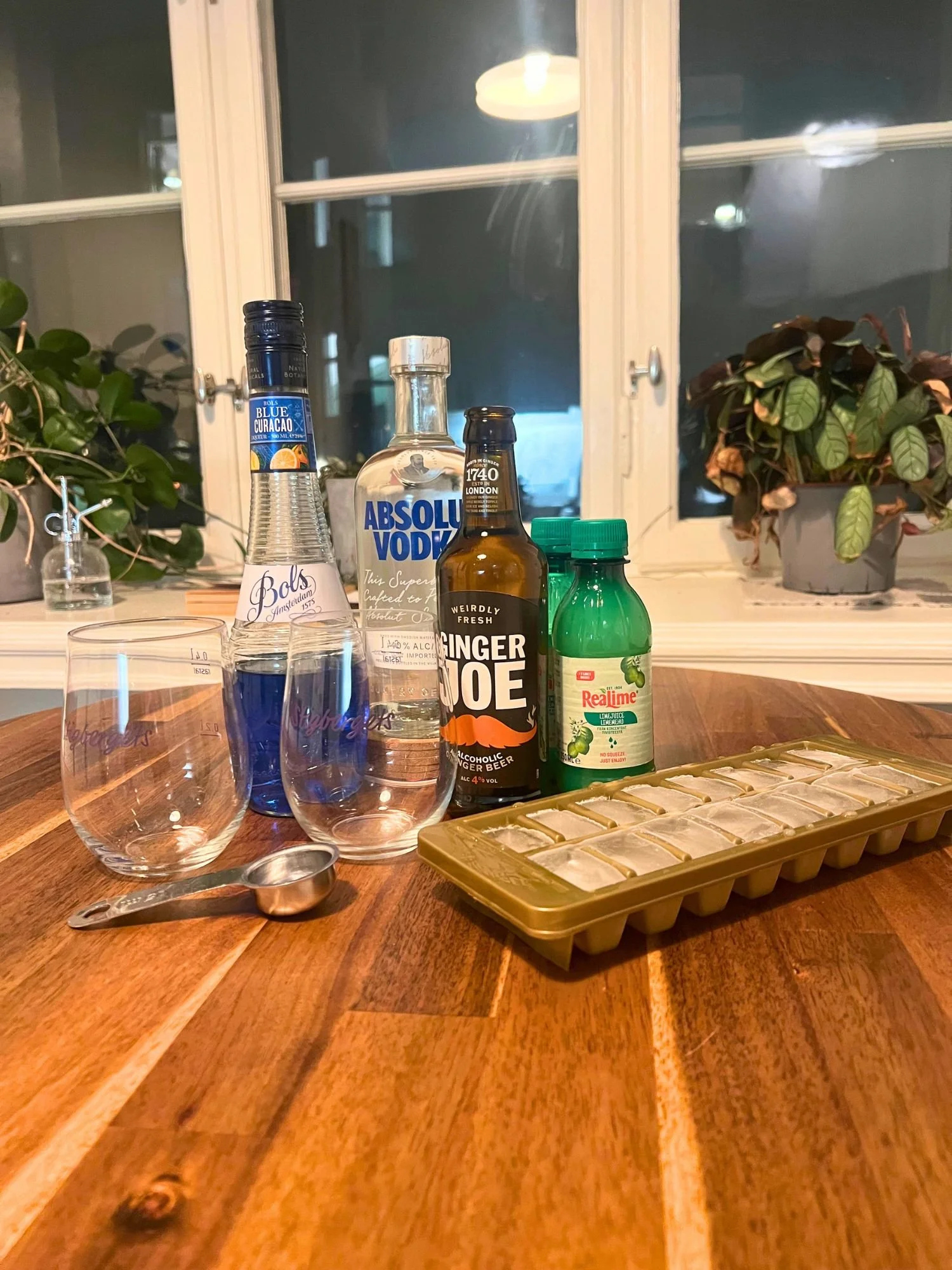 Ingredients for a blue cocktail on a wooden table, including vodka, blue curaçao, ginger beer, lime juice, ice cubes, measuring tools, and glasses, with a window and plants in the background.