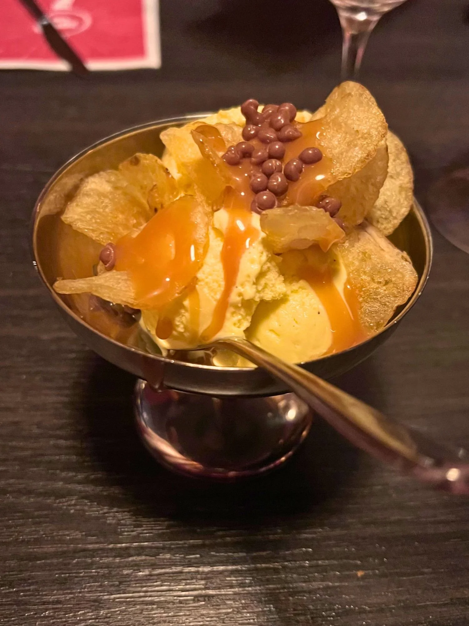 A metal dessert bowl on a dark table filled with vanilla ice cream, potato chips, a drizzle of caramel sauce, and small chocolate crispy pearls, served with a metal spoon.