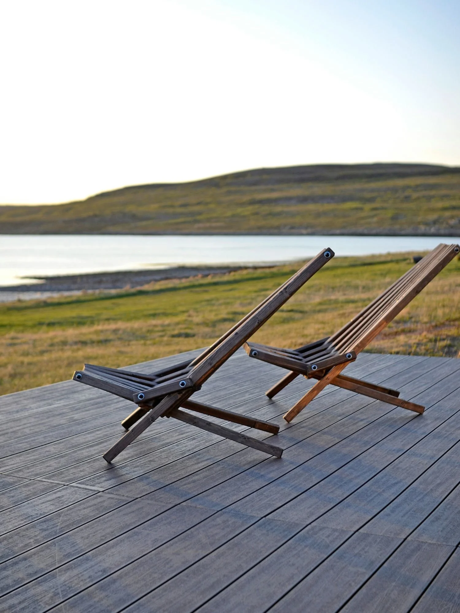 Two minimalist, slatted wooden lounge chairs sit empty on an outdoor wooden deck, positioned to look out over a scenic landscape.