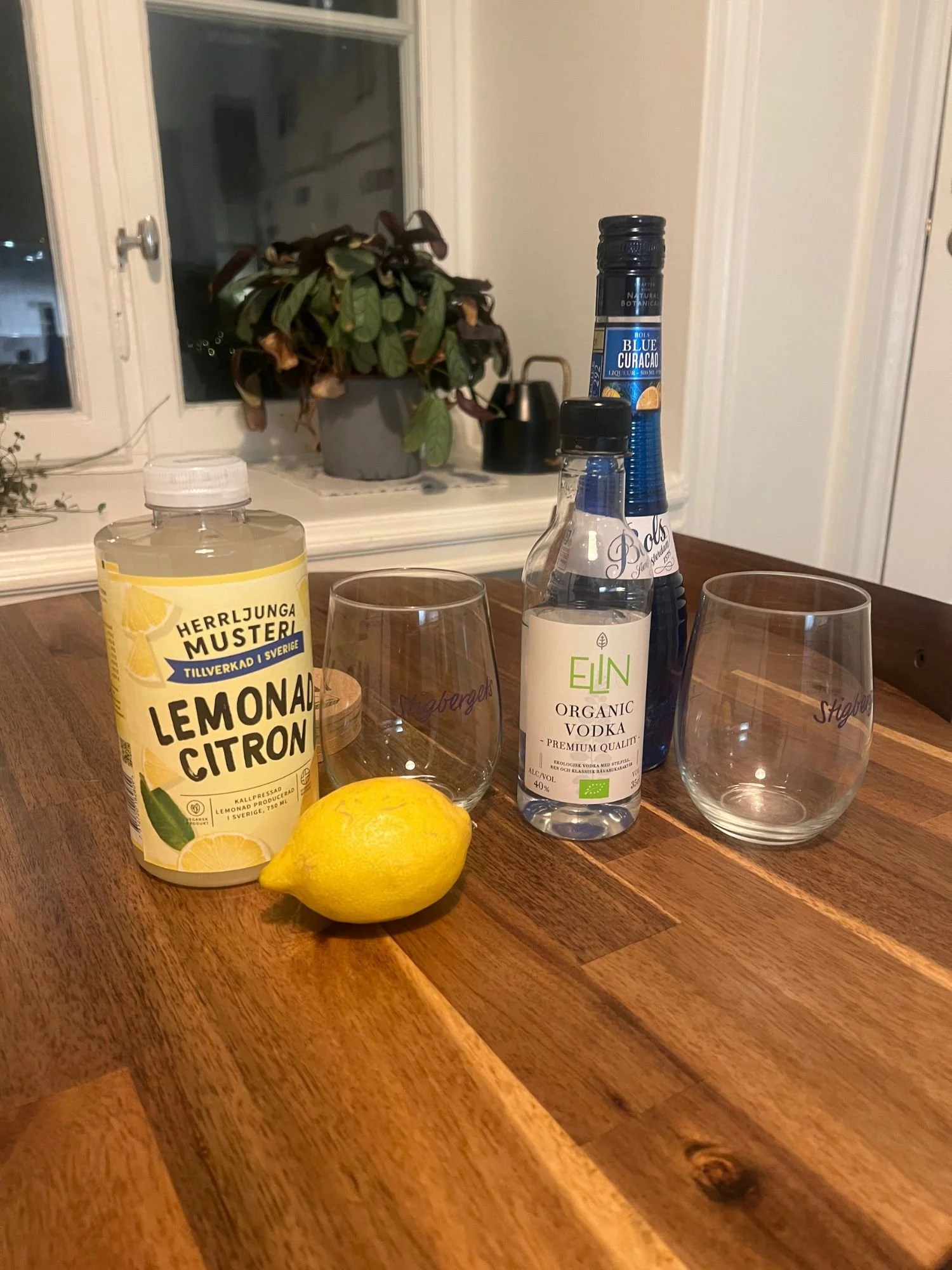 Ingredients for a blue cocktail on a wooden table, including lemonade, a lemon, vodka, blue curaçao, and two empty glasses, with a window and plants in the background.