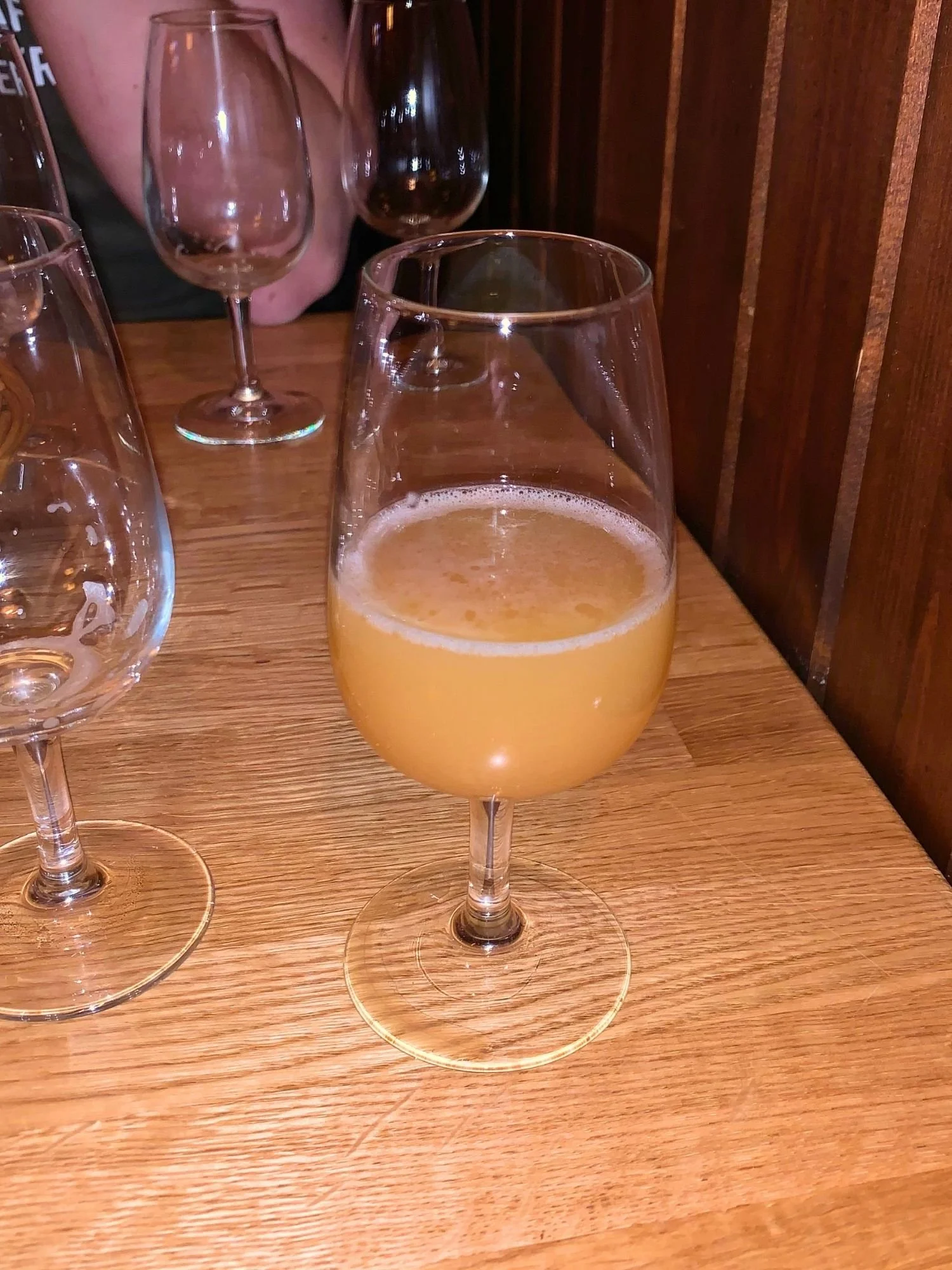 A hazy golden beer in a stemmed glass on a wooden table at a dimly lit bar, with several empty glasses in the background.