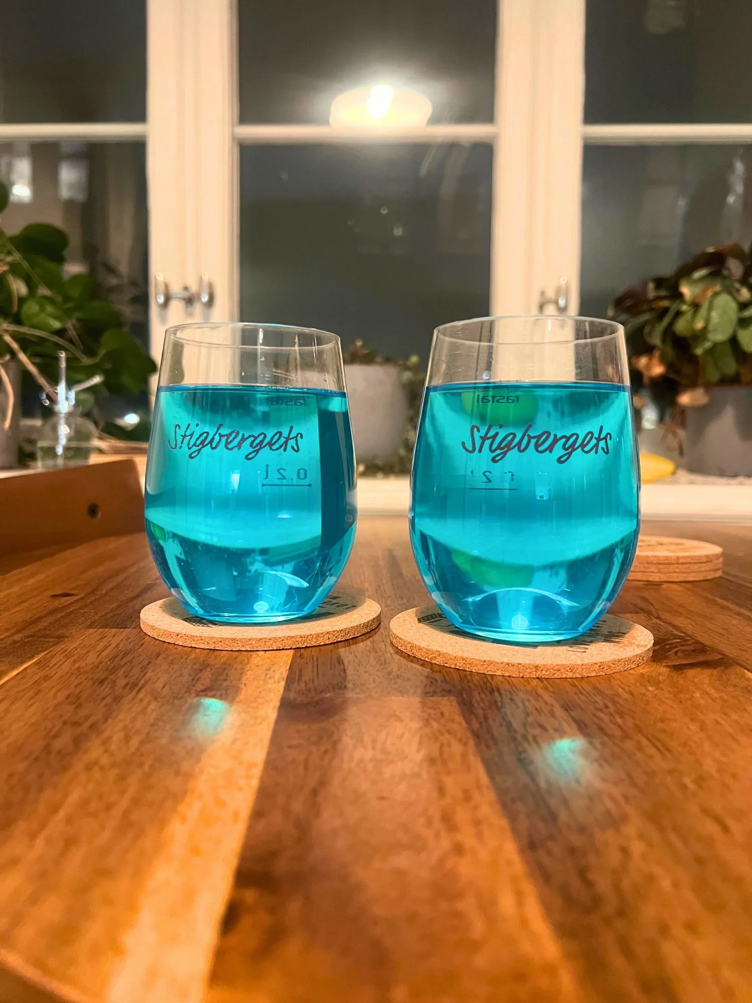 Two glasses of bright blue cocktails placed on coasters on a wooden table, with a window and houseplants in the background.
