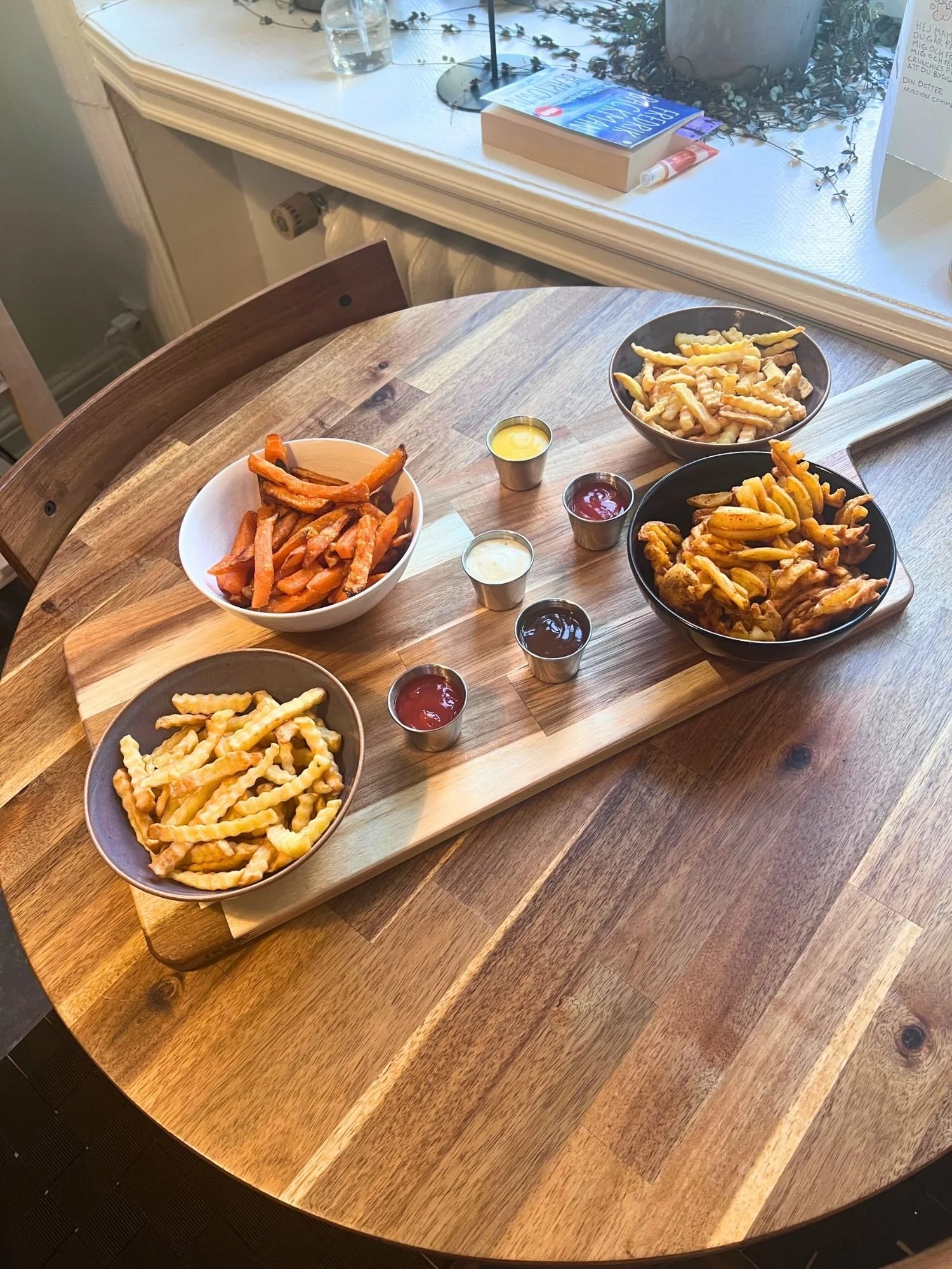 A wooden table set with a serving board featuring four bowls of different styles of fries and sweet potato fries, alongside five small metal cups of dipping sauces.