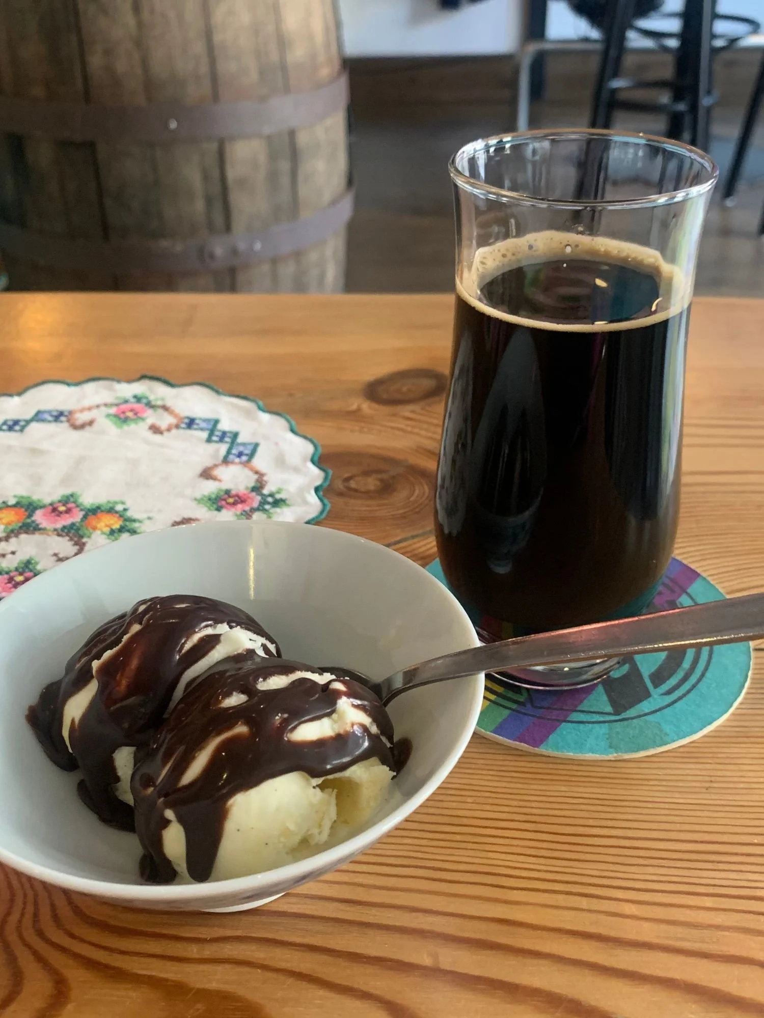 A small white ceramic bowl with two scoops of vanilla ice cream generously drizzled with a thick chocolate fudge sauce, with a metal spoon resting inside the bowl.