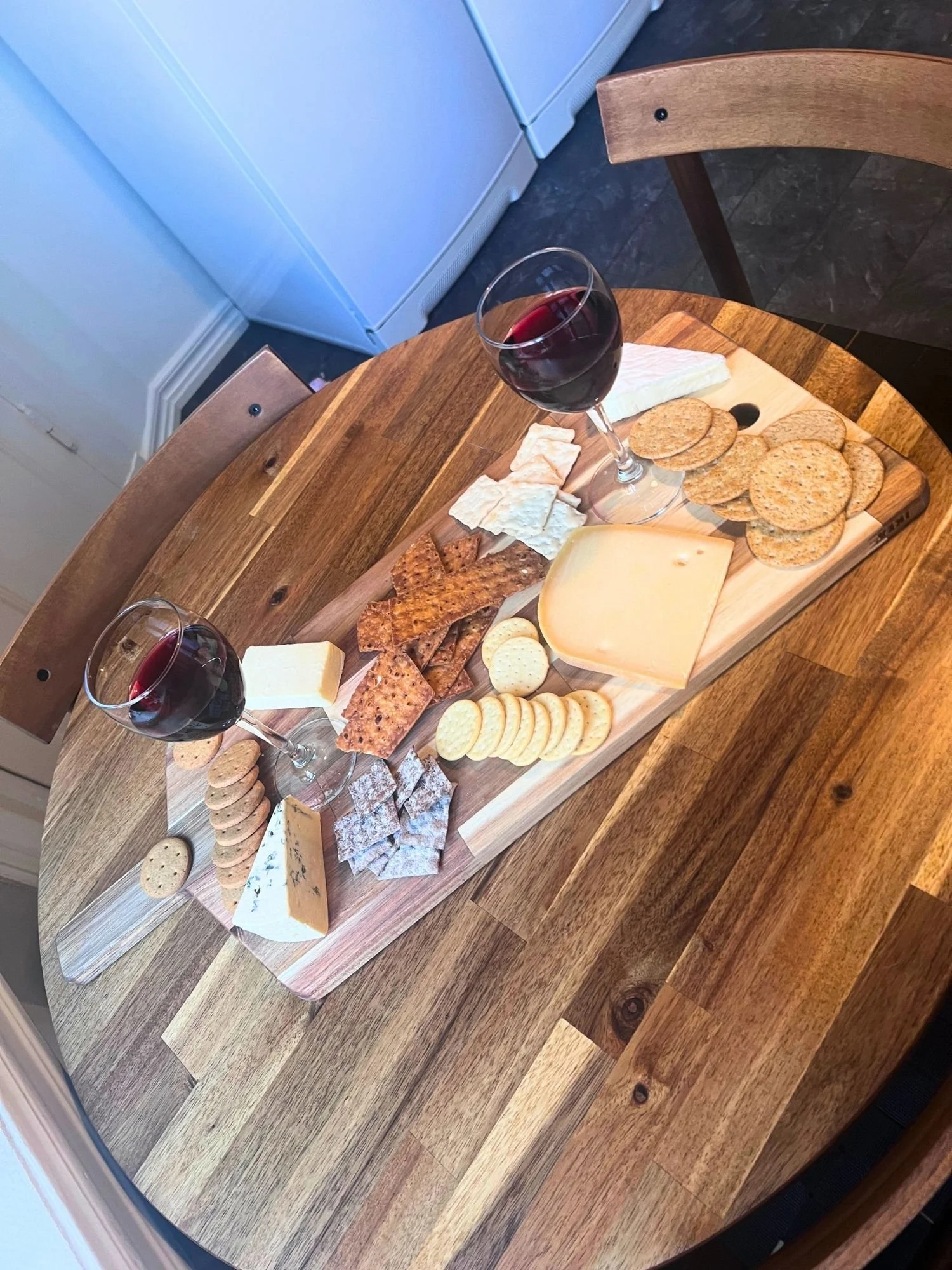 A wooden charcuterie board set diagonally on a round wooden table, arranged with various cheeses, assorted crackers, and two glasses of red wine.