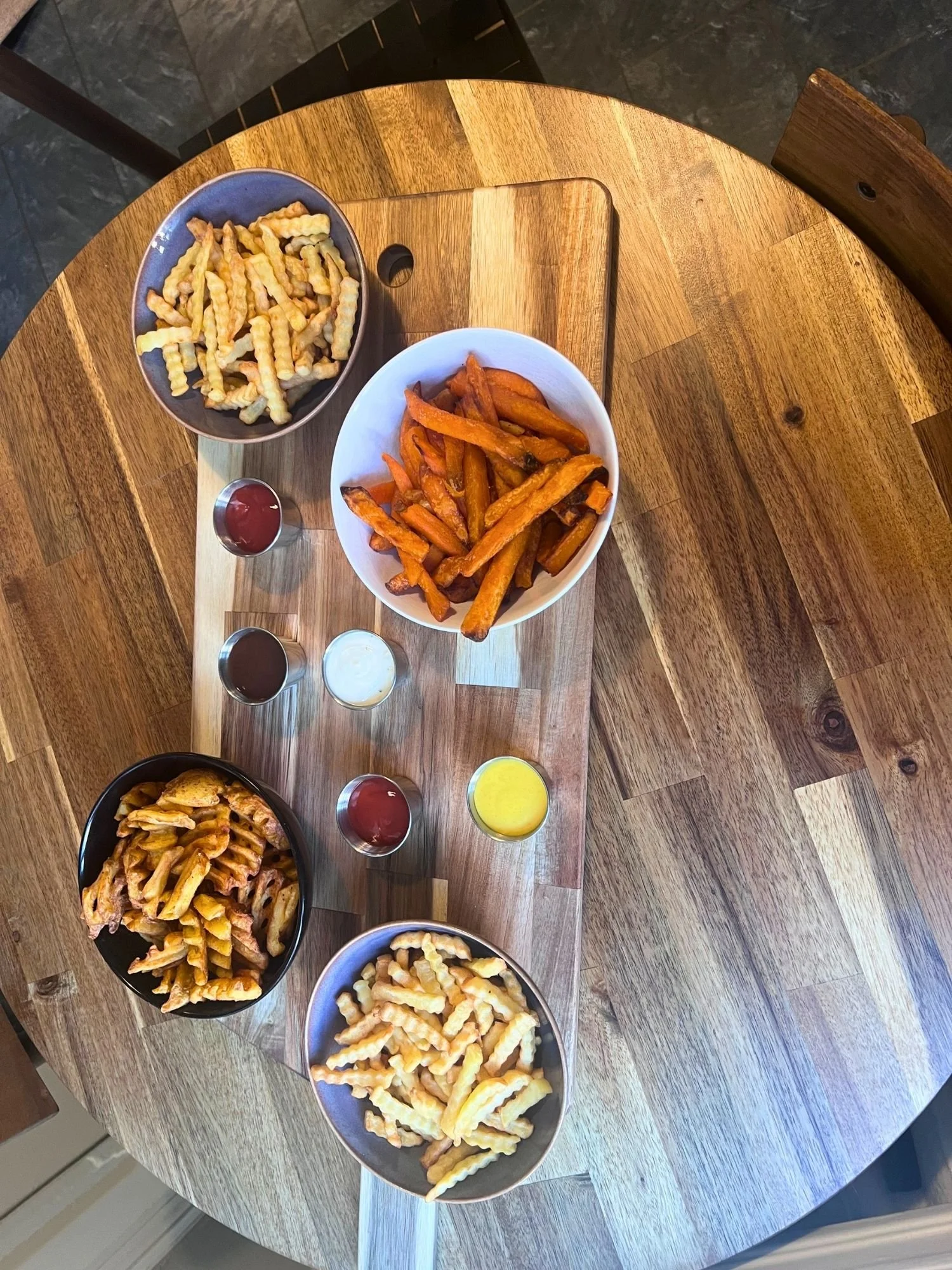 Overhead view of a wooden table set with a serving board holding four bowls of different styles of fries and sweet potato fries, alongside five small metal ramekins of dipping sauces.