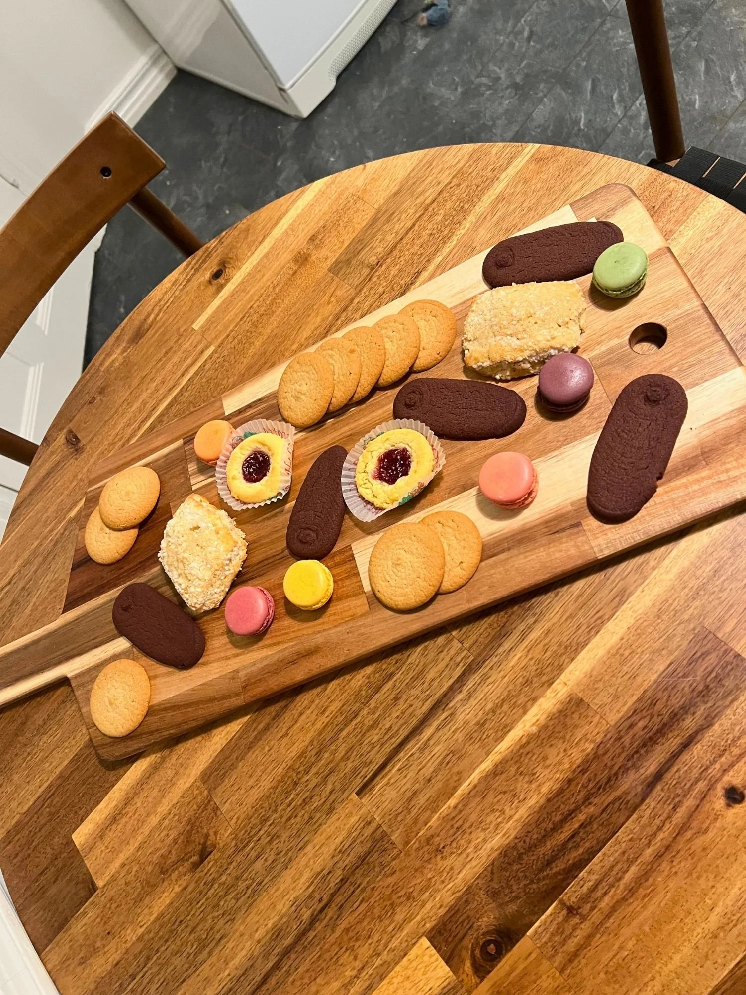 High-angle shot of a rectangular wooden serving board on a round wooden table, filled with an assortment of cookies, including colorful macarons, chocolate wafers, round butter cookies, jam-filled thumbprint cookies, and crusted pastries.