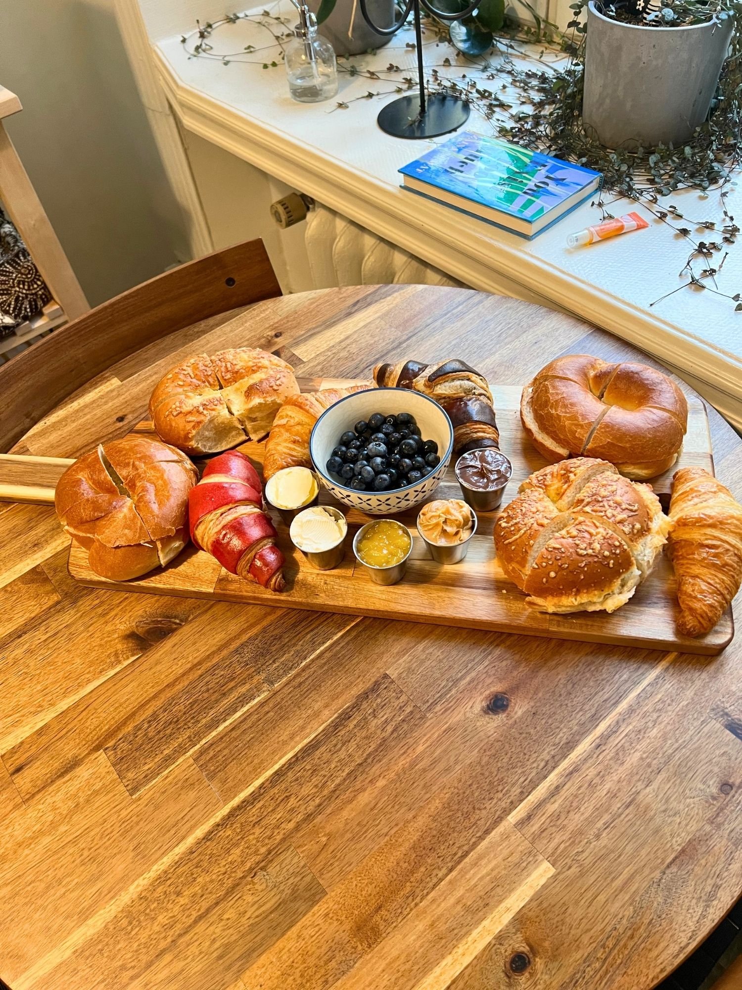 A wooden serving board on a round table filled with sliced bagels, assorted croissants, fresh blueberries, and small ramekins of sweet and savory spreads.