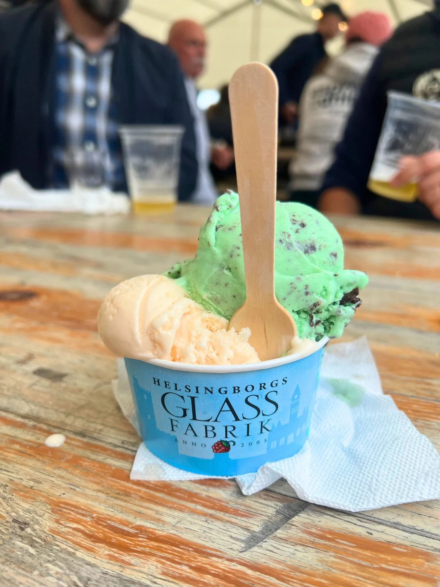A blue paper cup filled with two scoops of ice cream, one cookie dough and one mint chocolate chip, on a paper napkin on a wooden table. A wooden spoon is sticking upright out of the ice cream.