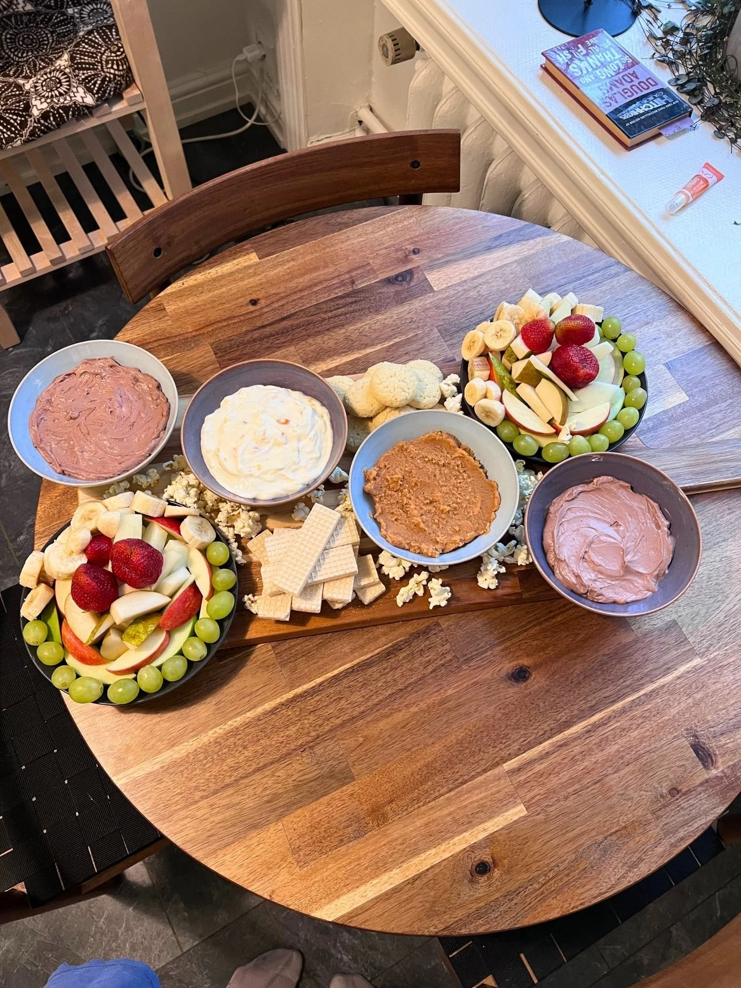 A wooden table set with a snack board holding two fruit platters, four ceramic bowls of dessert dips, light cookies, wafer biscuits, and scattered popcorn.