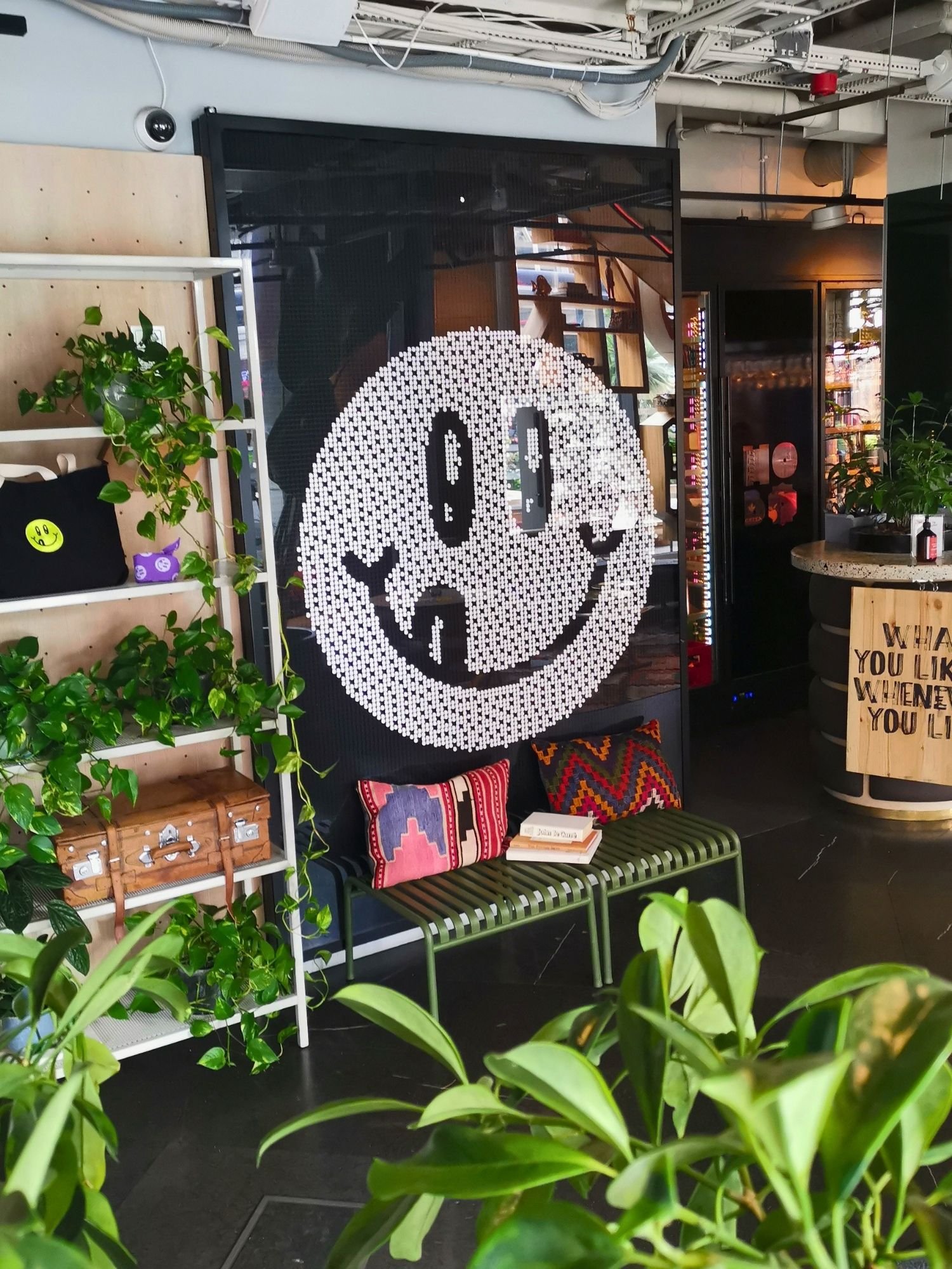 A large, white pixelated smiley face with a sticking-out tongue displayed on a dark glass wall inside a modern, plant-filled space. Below the wall sits a green metal bench styled with colorful patterned pillows and a small stack of books.