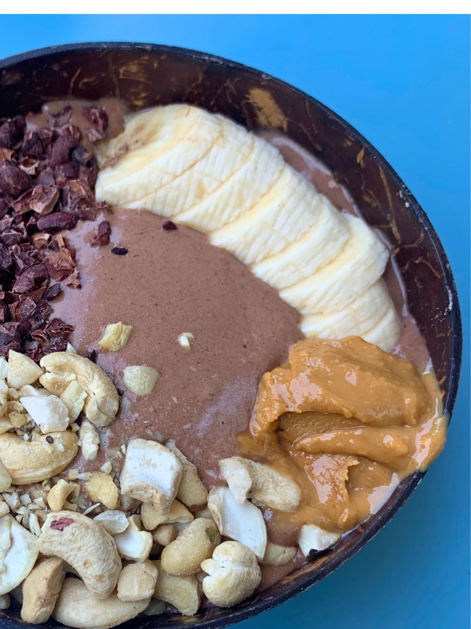 A chocolate smoothie bowl served in a dark bowl on a blue table. The bowl is neatly arranged with toppings including sliced bananas, a dollop of creamy peanut butter, whole and pieces of cashew nuts, and dark cacao nibs.