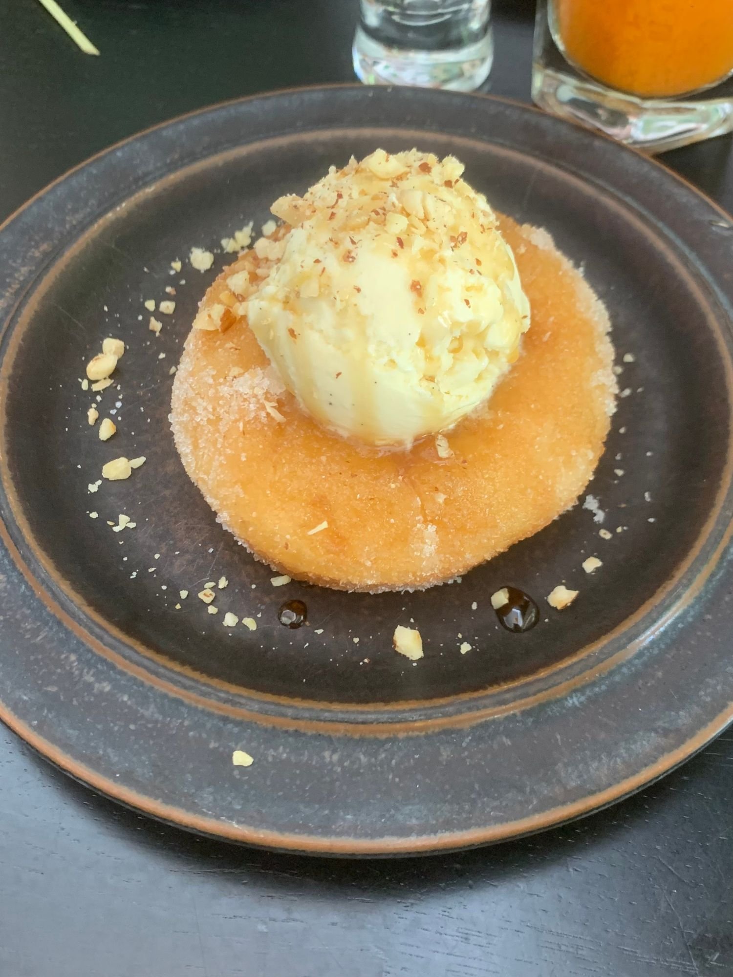 A sugar-dusted donut centered on a dark rimmed plate, topped with a scoop of vanilla ice cream, a drizzle of maple syrup, and chopped hazelnuts.