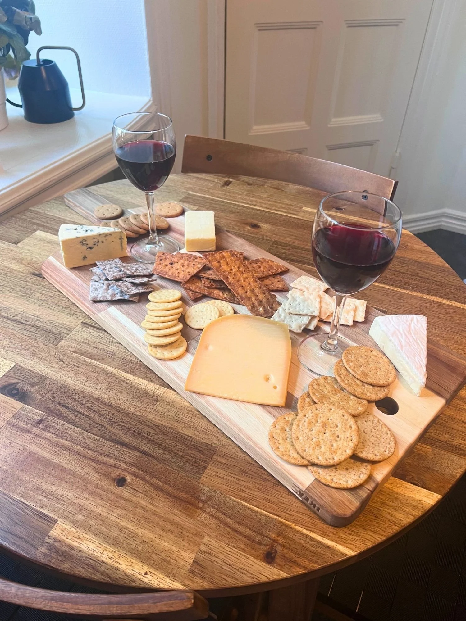 A wooden charcuterie board set on a round wooden table, filled with an assortment of cheeses, crackers, and two glasses of red wine in front of a white door.