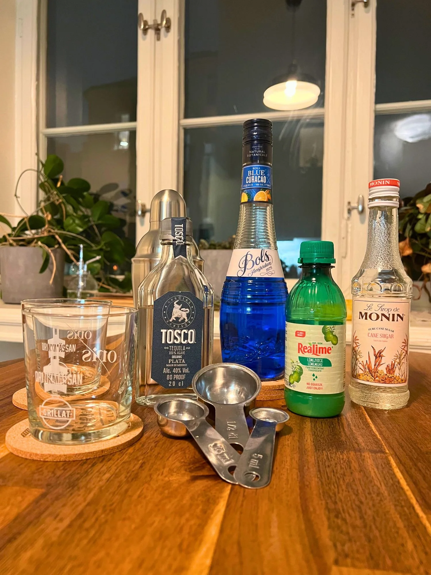 Ingredients for a blue cocktail on a wooden table, including tequila, blue curaçao, lime juice, simple syrup, a cocktail shaker, measuring spoons, and a glass, with a window and plants in the background.