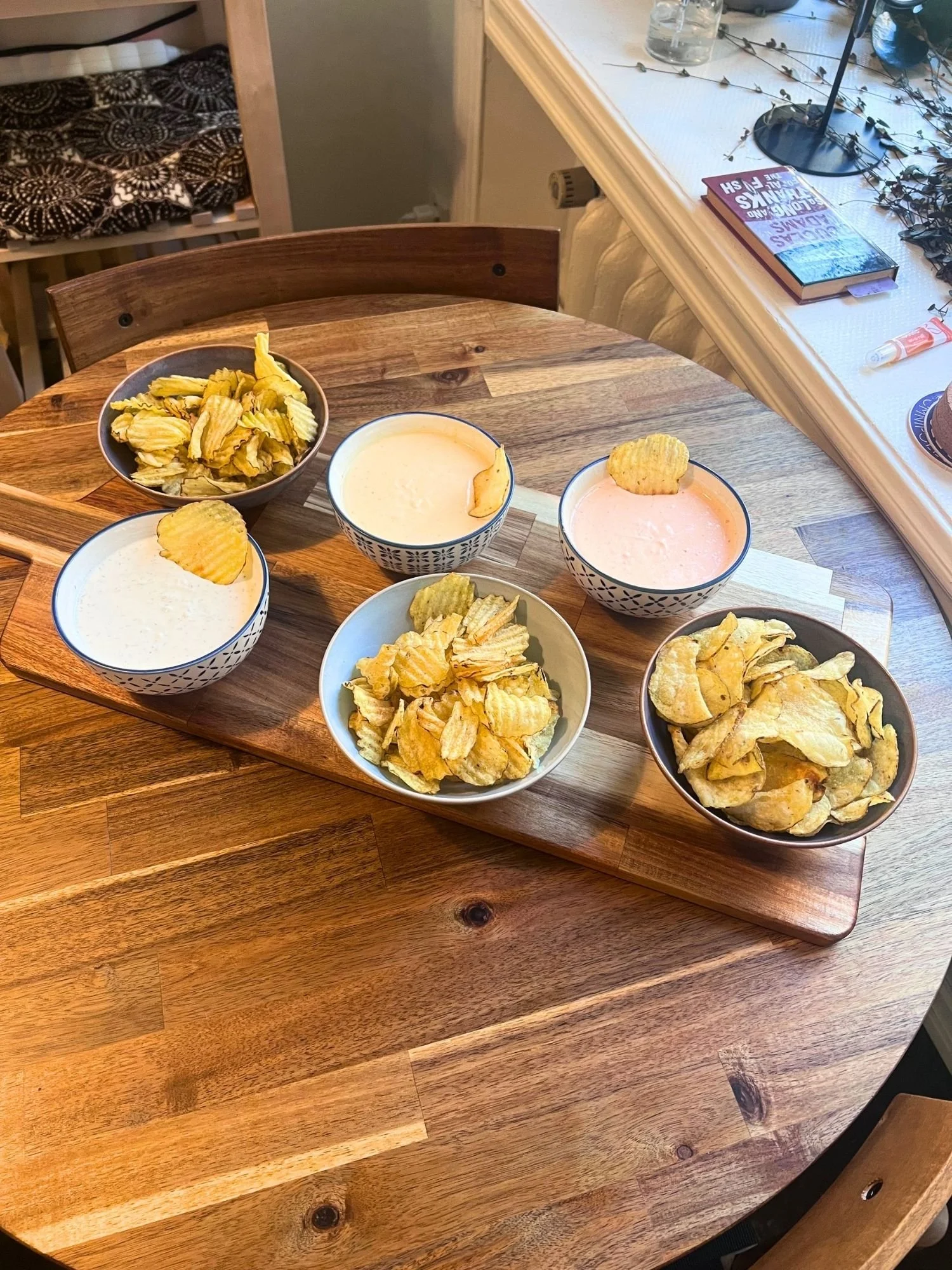 High-angle view of a wooden serving board set on a round wooden table, featuring three bowls of potato chips and three bowls of creamy dips.