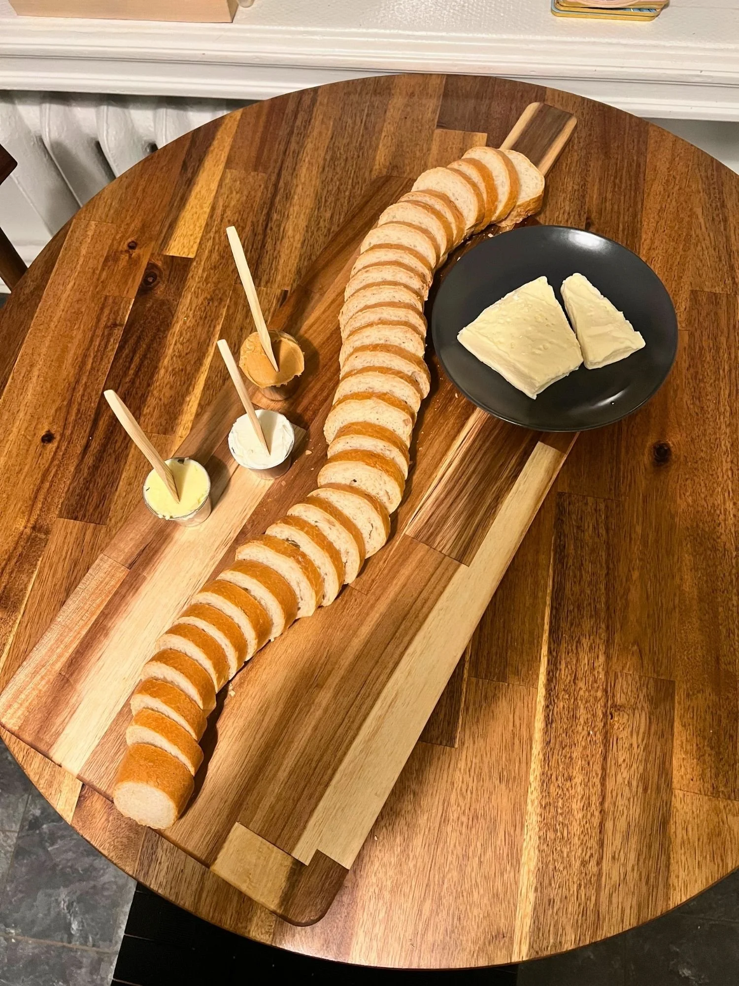 A long wooden serving board on a round wooden table features a whole sliced baguette arranged in an "S" curve down the length of the board.
