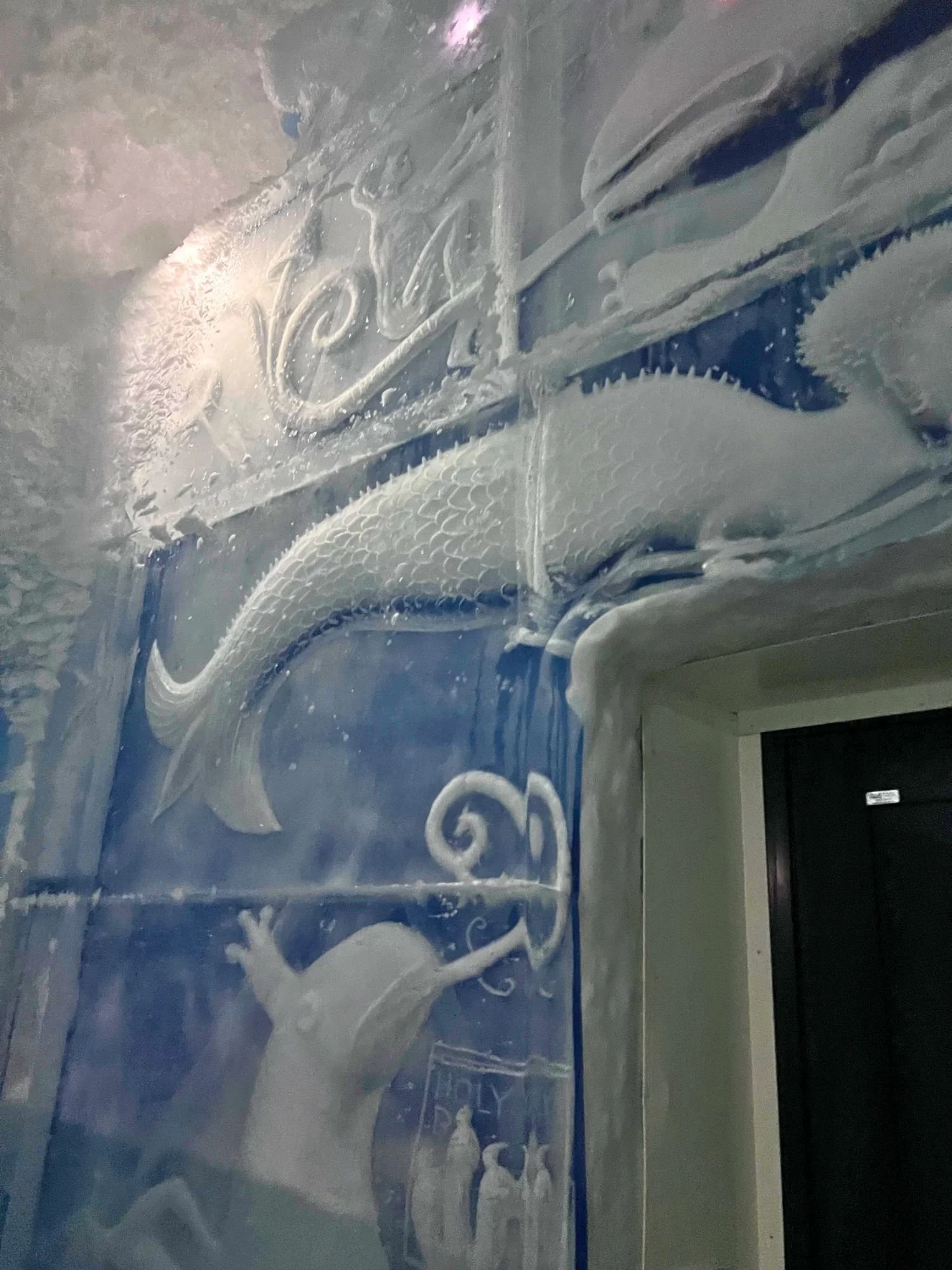 a large ice wall carving next to a doorway inside a room at the Icehotel.  The blue-tinted, translucent ice wall features several mythical and prehistoric sea creatures.
